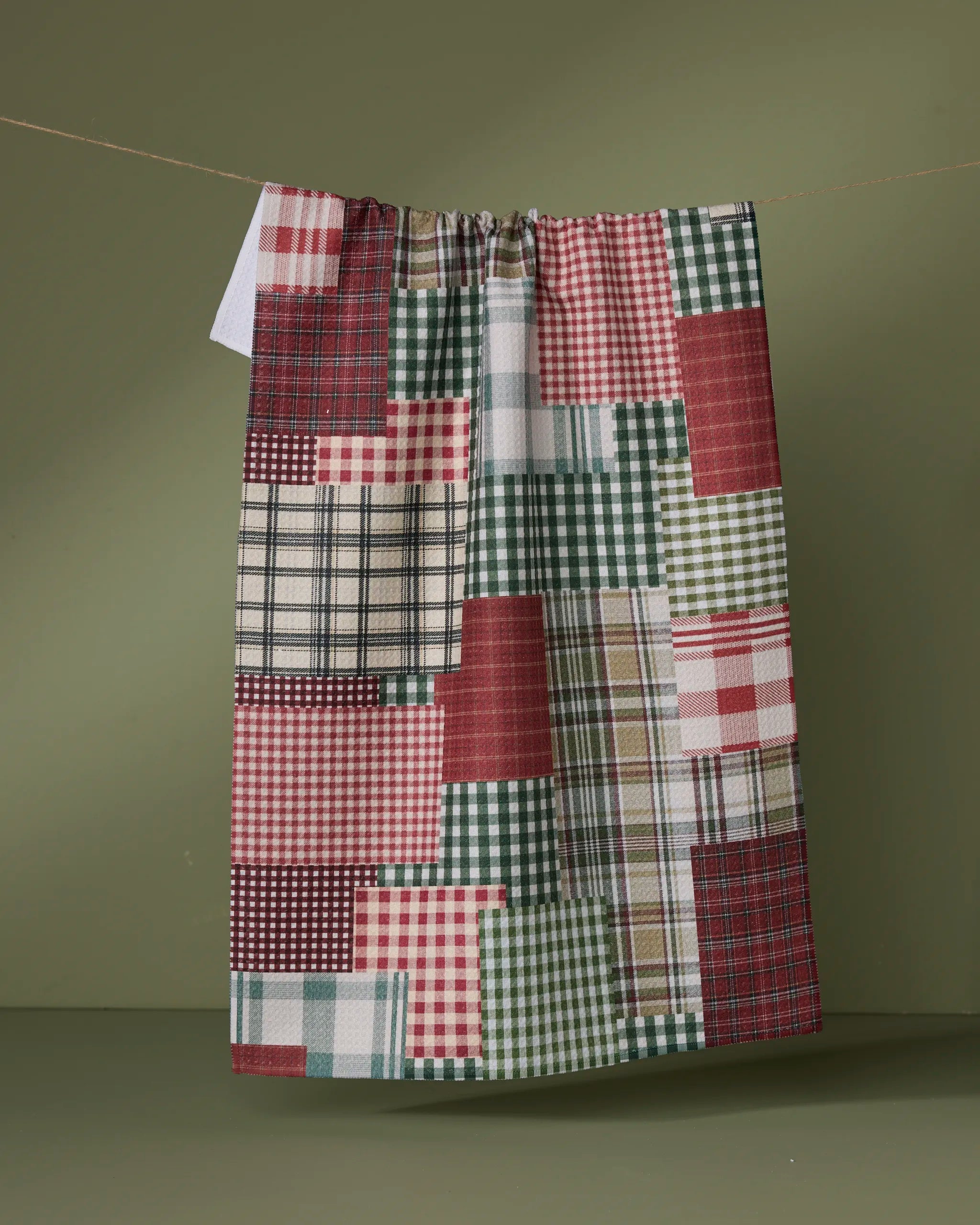 The Geometry Patchly tea towel, featuring ultra-absorbent fabric and a red, green, beige, and white plaid patchwork design, hangs on a line against an olive green background.