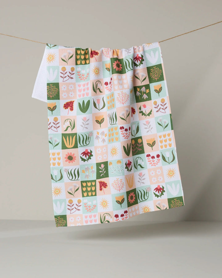 The "Patchwork Garden" tea towel by Rachel Parker features colorful floral and plant patchwork in green, yellow, pink, and red on recycled fabric, shown hanging on a clothesline against a neutral background.