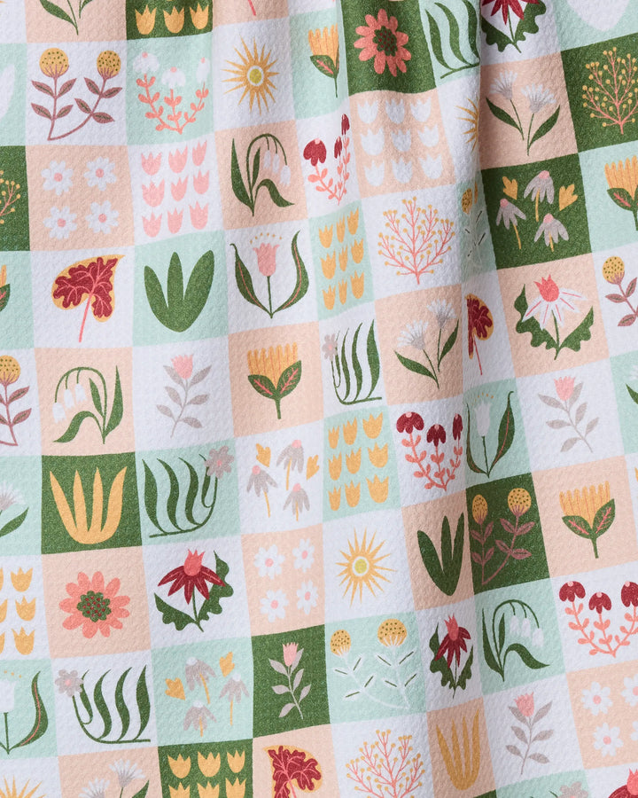 The Patchwork Garden tea towel by Rachel Parker features a grid of colorful flowers, leaves, and suns in green, pink, yellow, and red. Softly draped fabric creates gentle folds for a vibrant, textured look.
