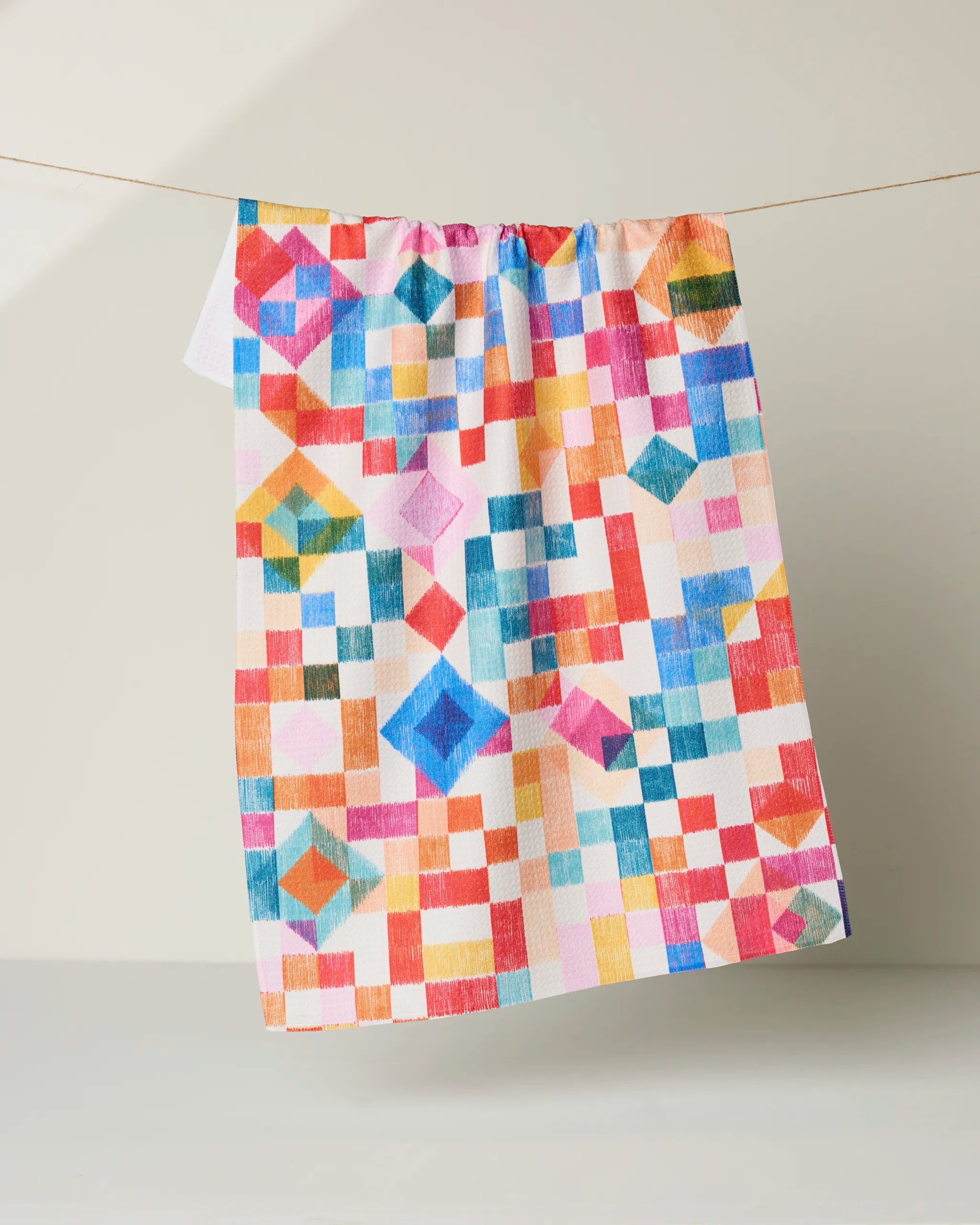 The Patchwork Geo tea towel by Rachel Parker features a vibrant geometric pixel design in red, blue, yellow, orange, and pink. Made from quick-drying recycled materials, it hangs on a line against a neutral background.