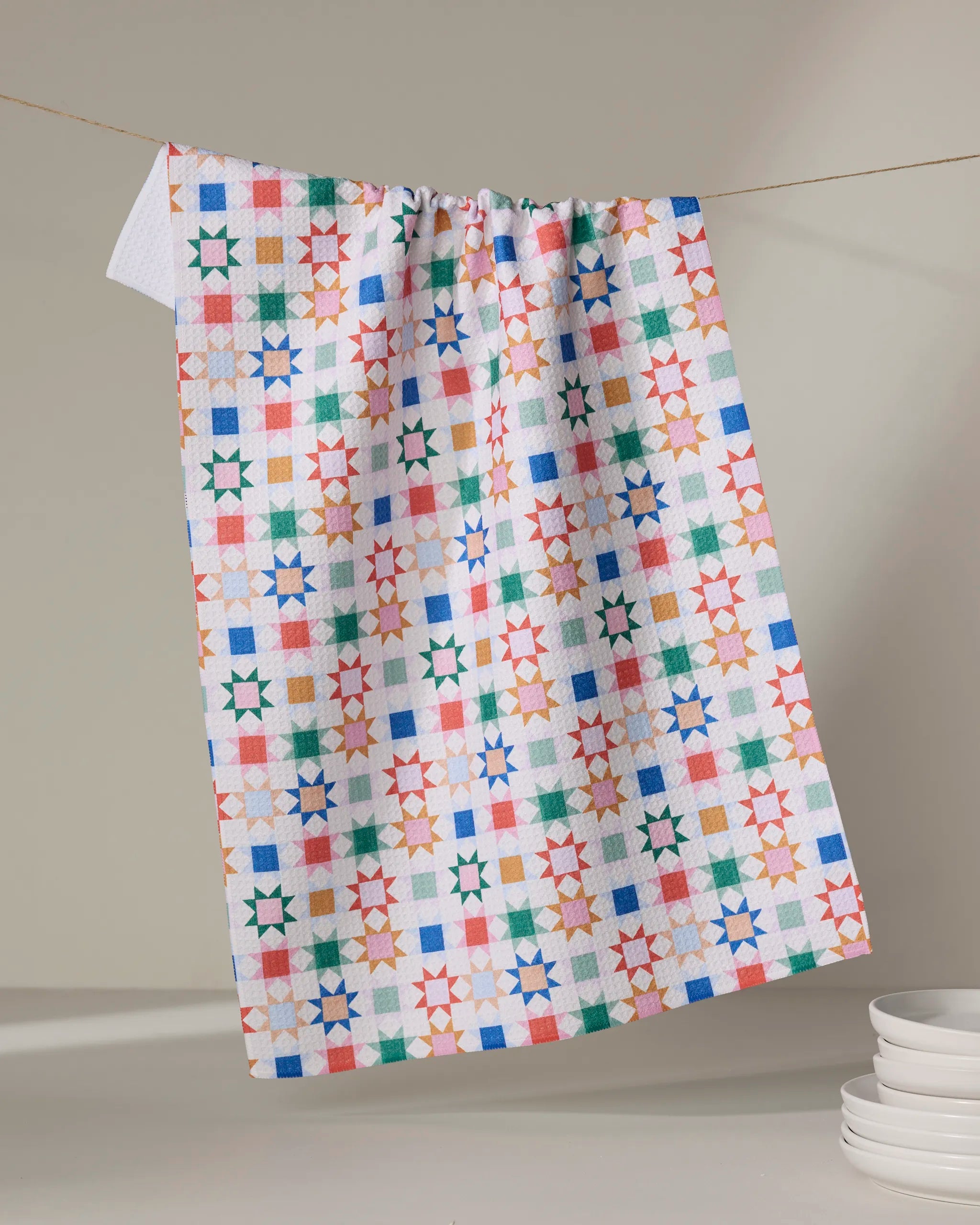 The Patchwork Picnic kitchen towel by Brittany Jepsen, featuring a vibrant geometric star and square design, hangs above a stack of white dishes against a neutral background.