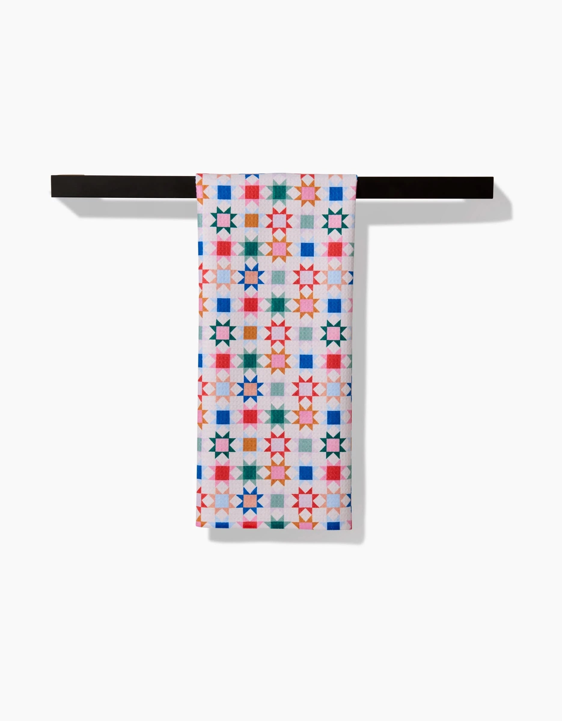 The Patchwork Picnic tea towel by Brittany Jepsen, crafted from Geoweave fabric, displays a vibrant geometric pattern of multicolored stars and squares, neatly folded over a black bar on a white background.