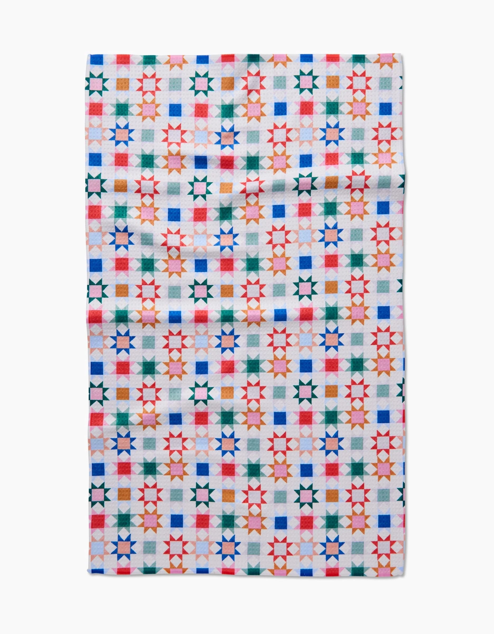 The Patchwork Picnic tea towel by Brittany Jepsen is made from Geoweave fabric and features a vibrant geometric grid of red, green, blue, and orange stars and squares on a white background.