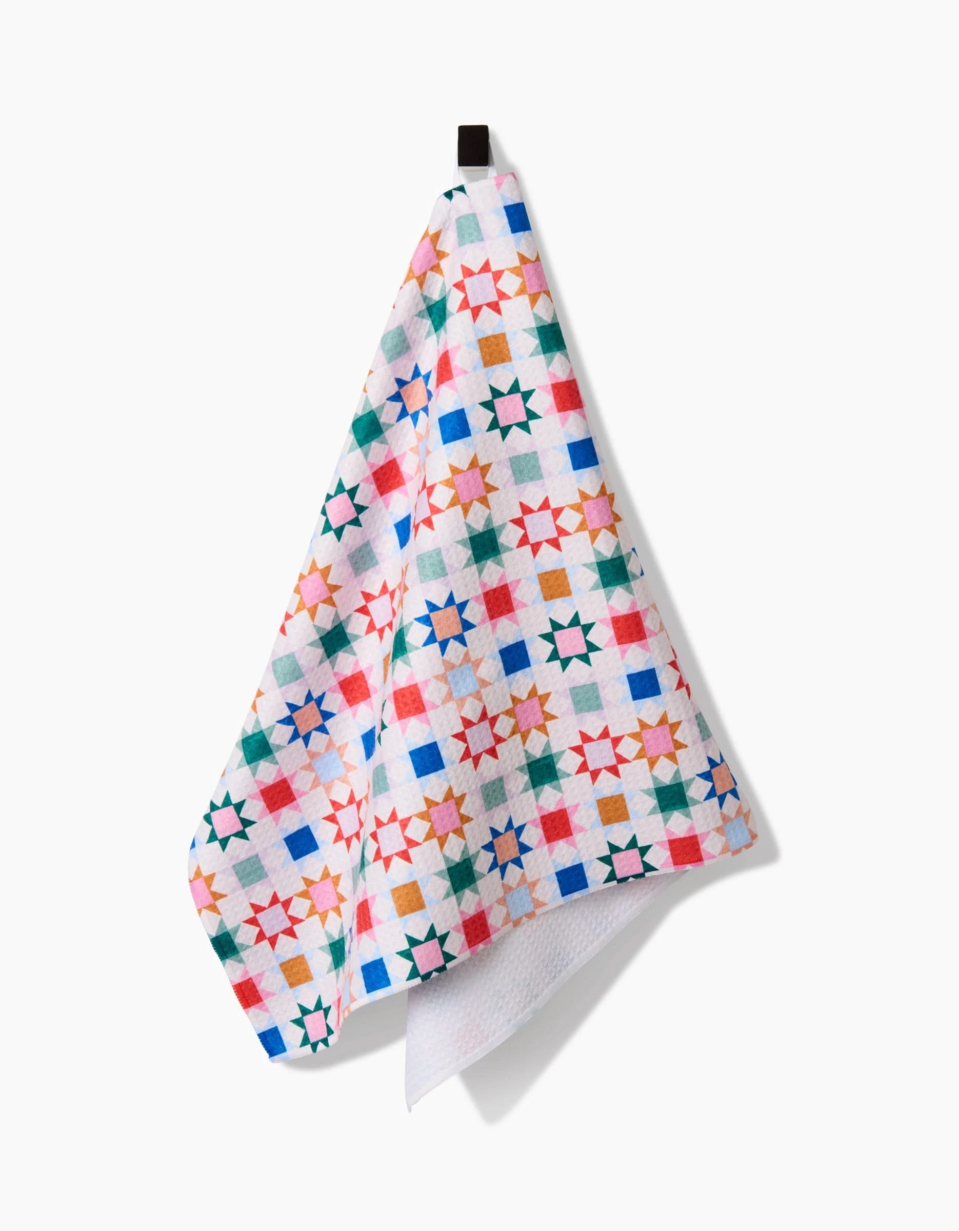 The Patchwork Picnic tea towel by Brittany Jepsen features a vibrant geometric star and square pattern in red, blue, green, orange, and pink on Geoweave fabric, shown hanging from a hook against a white background.
