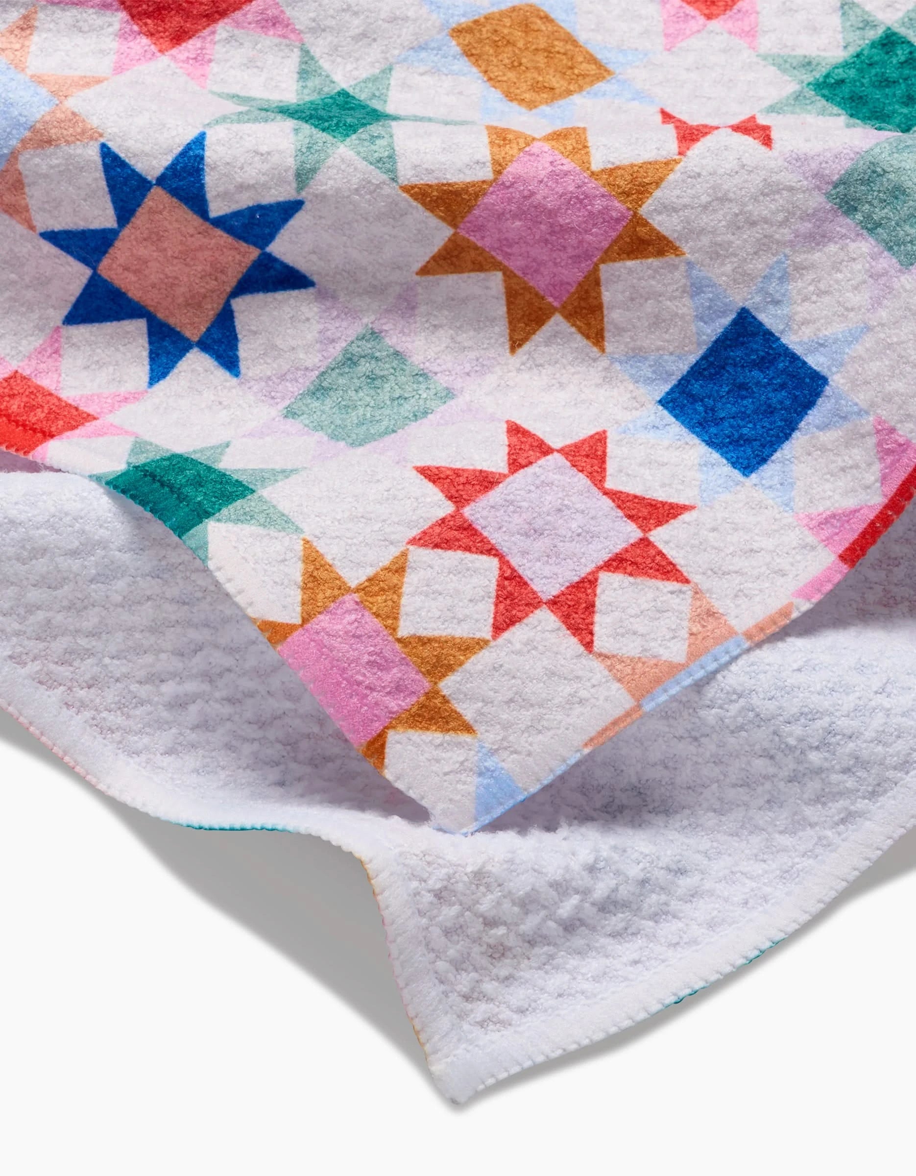 The “Patchwork Picnic” tea towel by Brittany Jepsen features a geometric quilt-inspired pattern with colorful star shapes on a white background, made from soft Geoweave fabric and shown with one corner folded to reveal the underside.