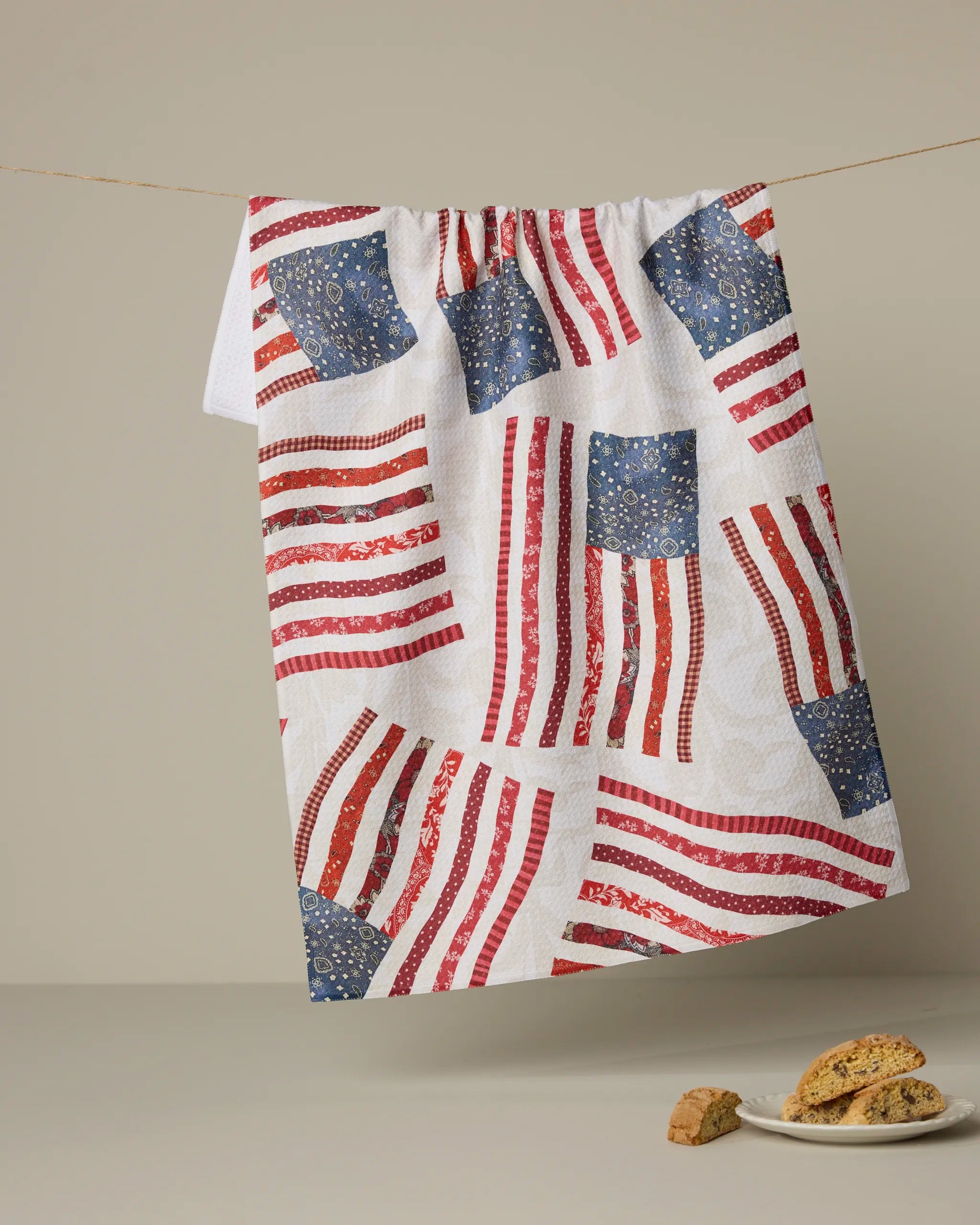 The Patchwork Pride tea towel by Geometry, featuring red and blue stylized American flags, hangs from a string, with several pieces of biscotti on the surface below.