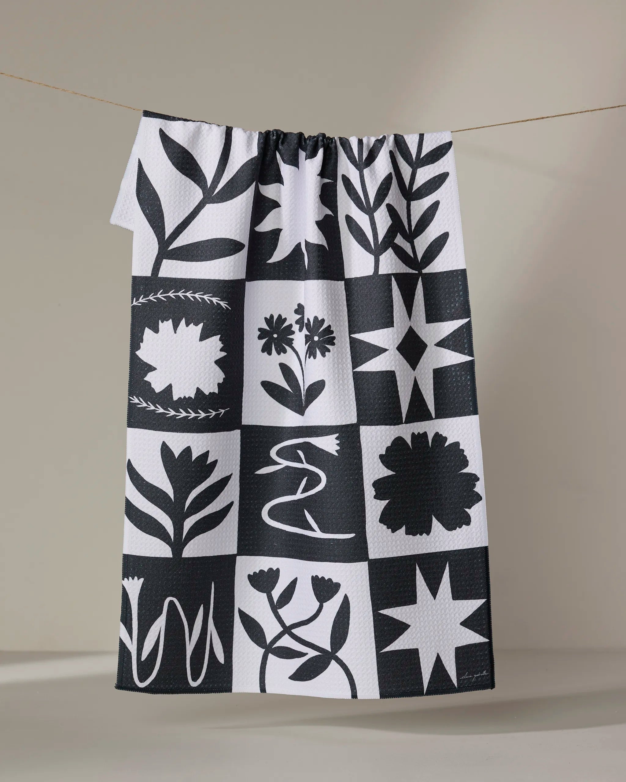 The Patchwork kitchen towel by Elana Gabrielle, made from recycled materials, features a black and white grid pattern with bold floral and geometric designs, hanging on a line against a neutral background.