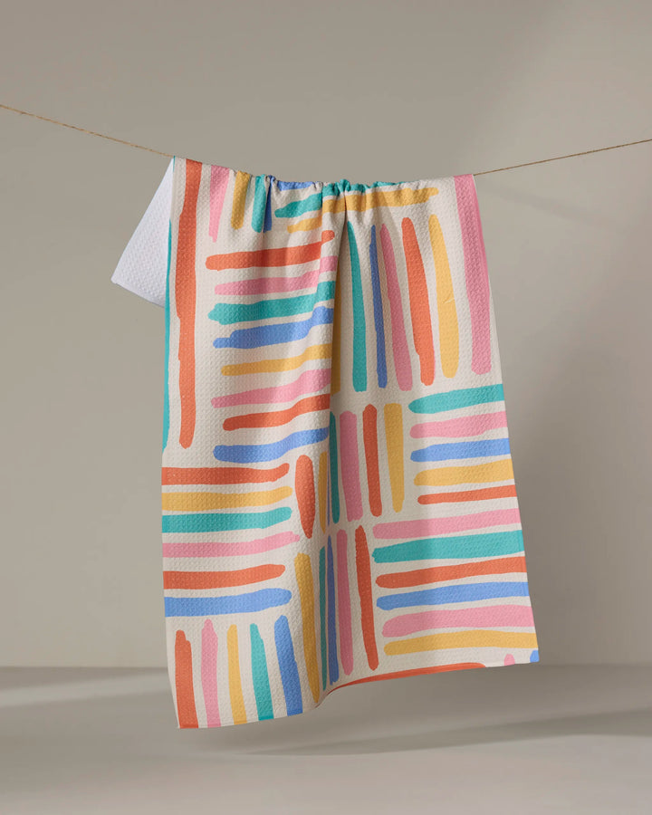 The Pathways tea towel by Sareka Unique features vibrant pink, orange, blue, and green brushstrokes in a grid pattern and hangs on a string against a neutral background.