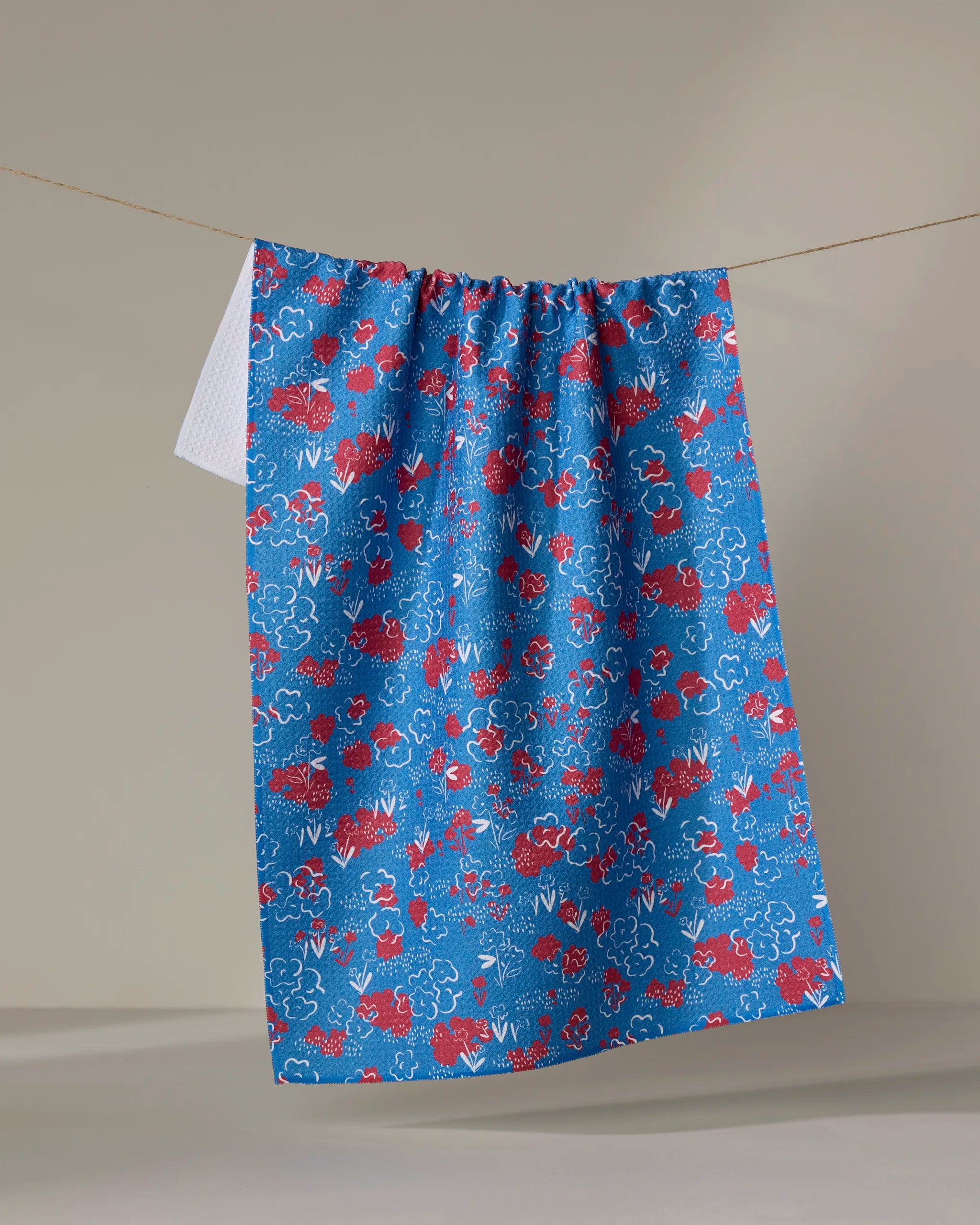 The Patriotic Garden kitchen towel by Second Nature Studio, featuring a red and white floral pattern on blue recycled fabric, hangs on a clothesline with a second white towel partly visible against a plain background.