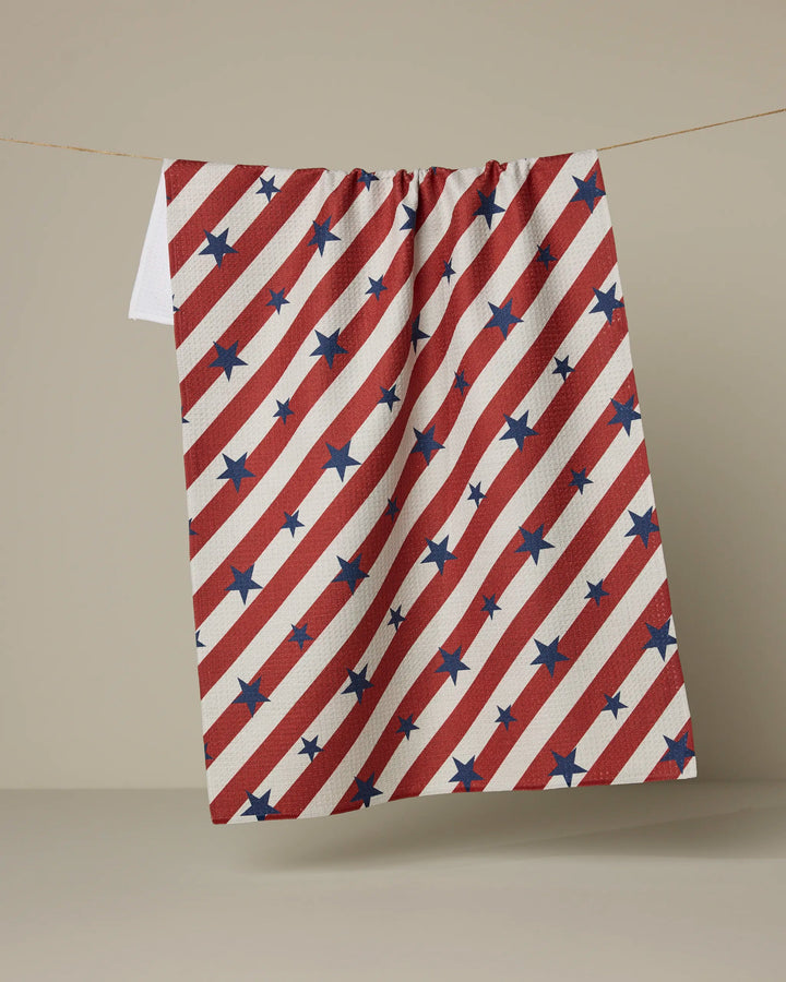 The Geometry "Patriotic Stripe" kitchen towel, featuring diagonal red and white stripes with blue stars, hangs on a clothesline against a neutral background.
