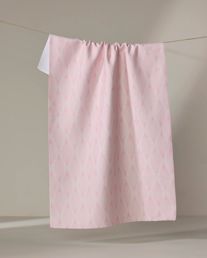 The Shelley Smith "Patterned in Pink" kitchen towel, made from recycled materials with a subtle diamond design, hangs neatly on a clothesline against a neutral background.