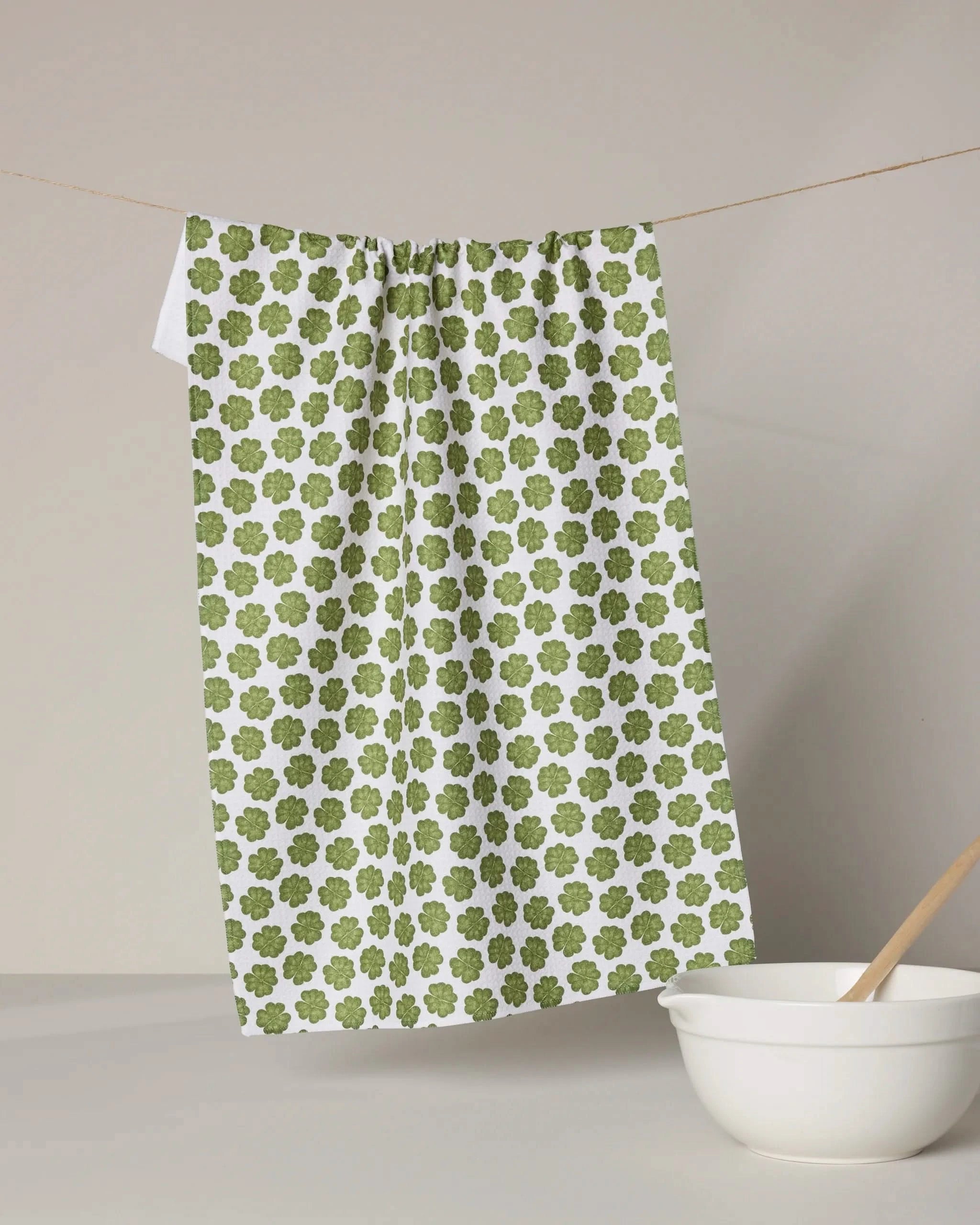 The Geometry Patty tea towel, featuring a green clover print on white, hangs above a white countertop, while a white mixing bowl and wooden spoon rest in the foreground.