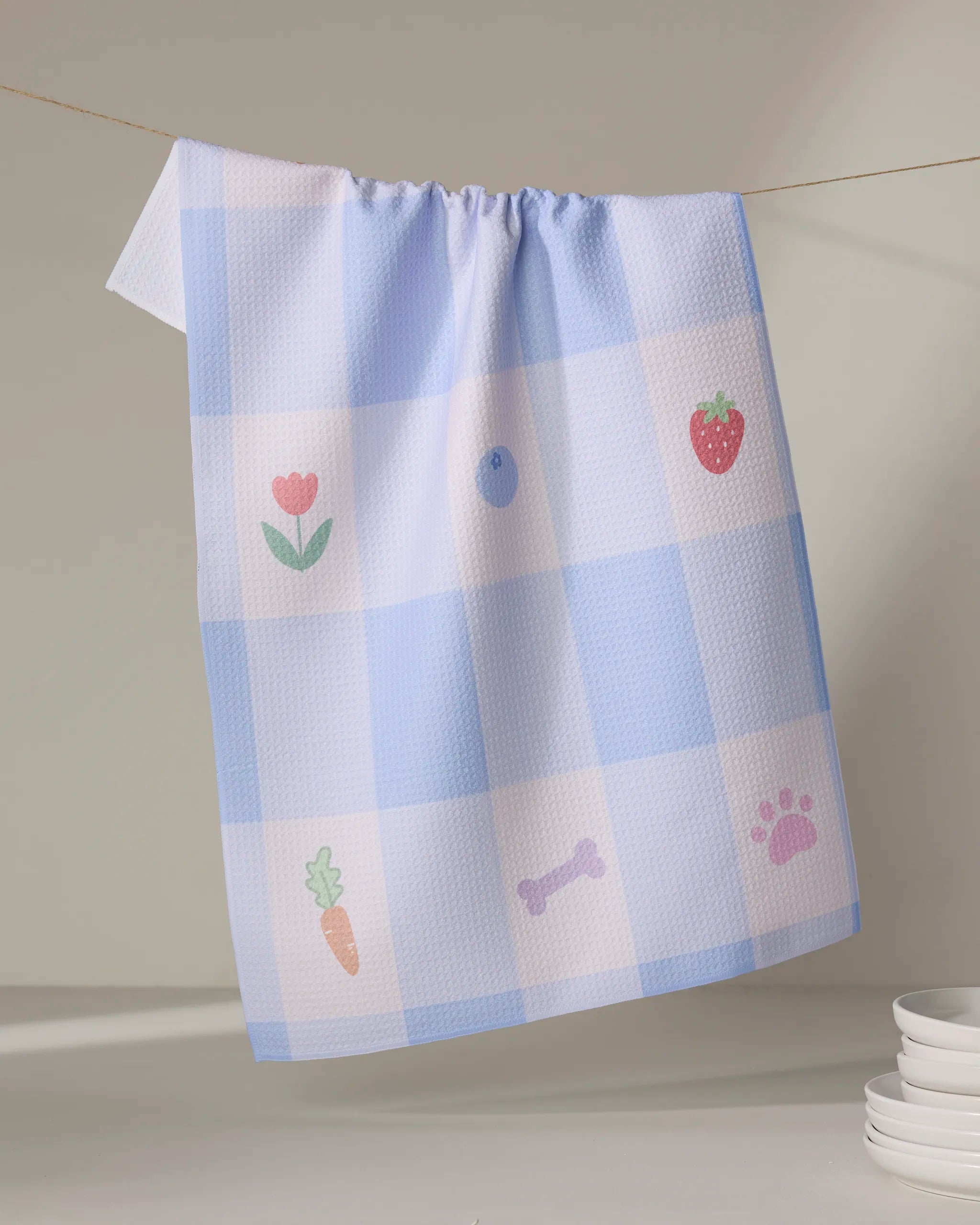 The “Pawfect Picnic” tea towel by Letters By Shells, with blue and white checks and cute icons like tulips, blueberries, and paw prints, hangs near a stack of white plates—bringing whimsy to your kitchen.
