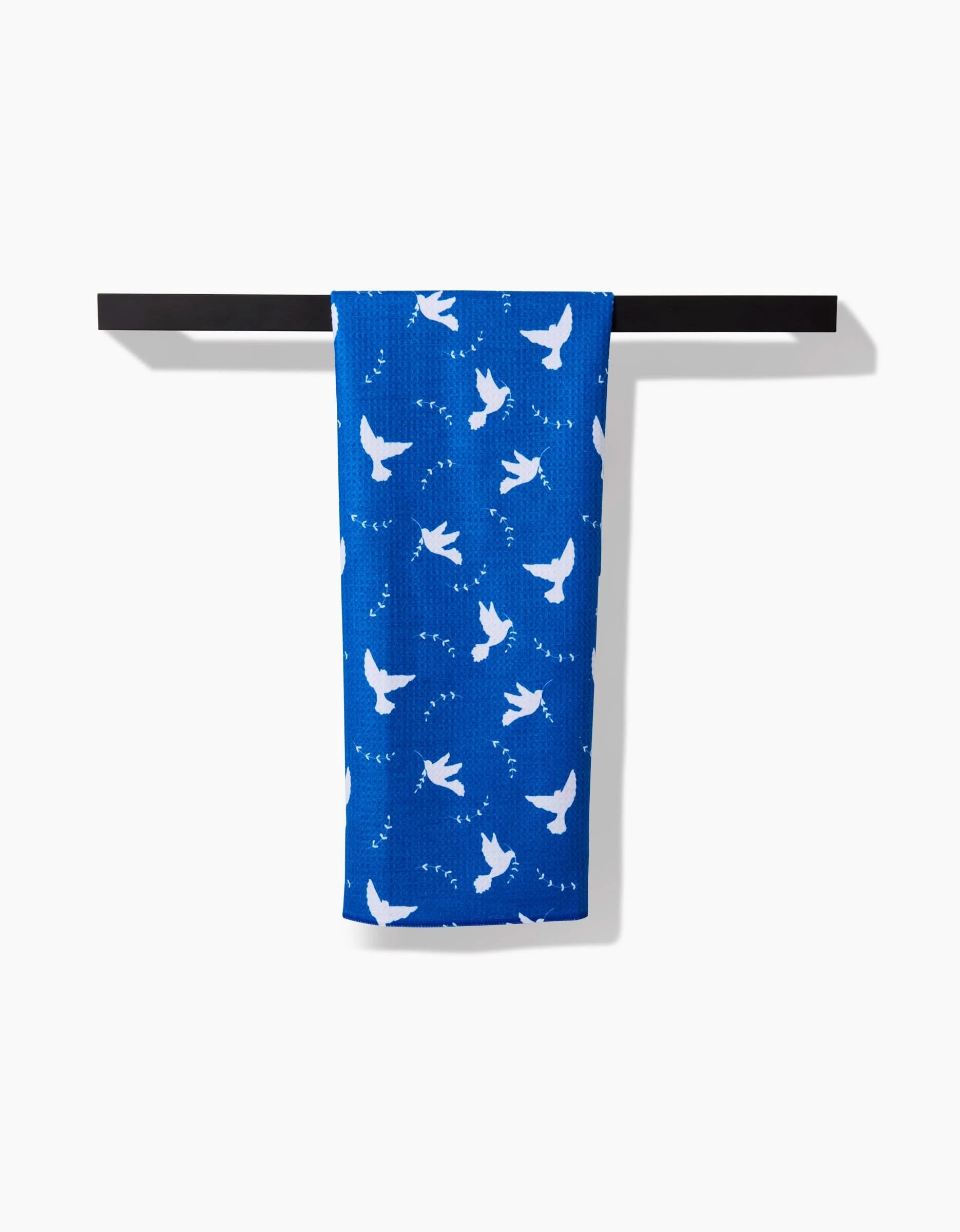 The Peace Dove tea towel by Pascale Dilger, made from recycled materials and adorned with a white bird pattern, hangs on a sleek black towel bar against a white background.