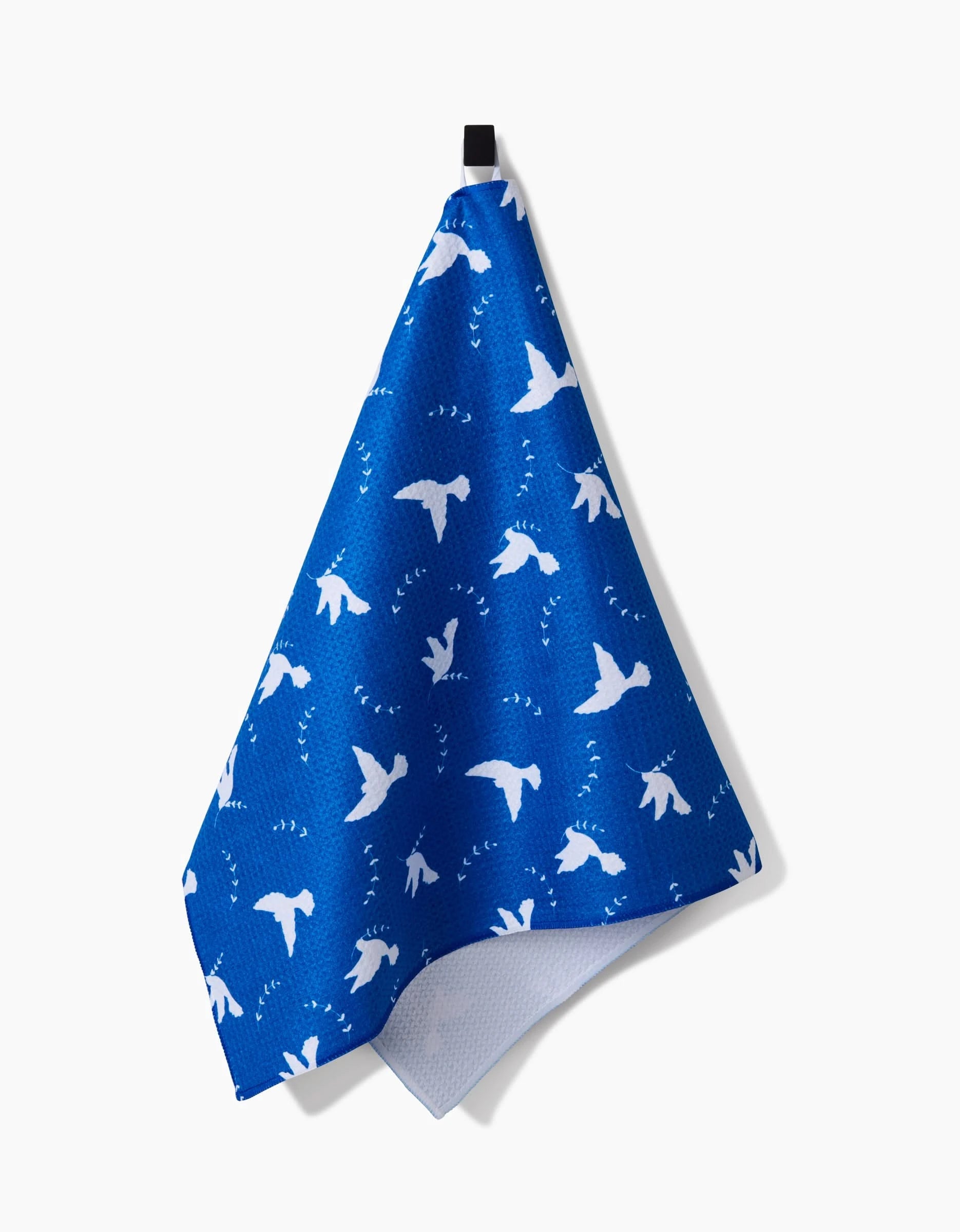 The Pascale Dilger Peace Dove kitchen towel, made from recycled materials, features white dove silhouettes and small flying shapes on blue fabric and hangs from a black loop against a plain white background.