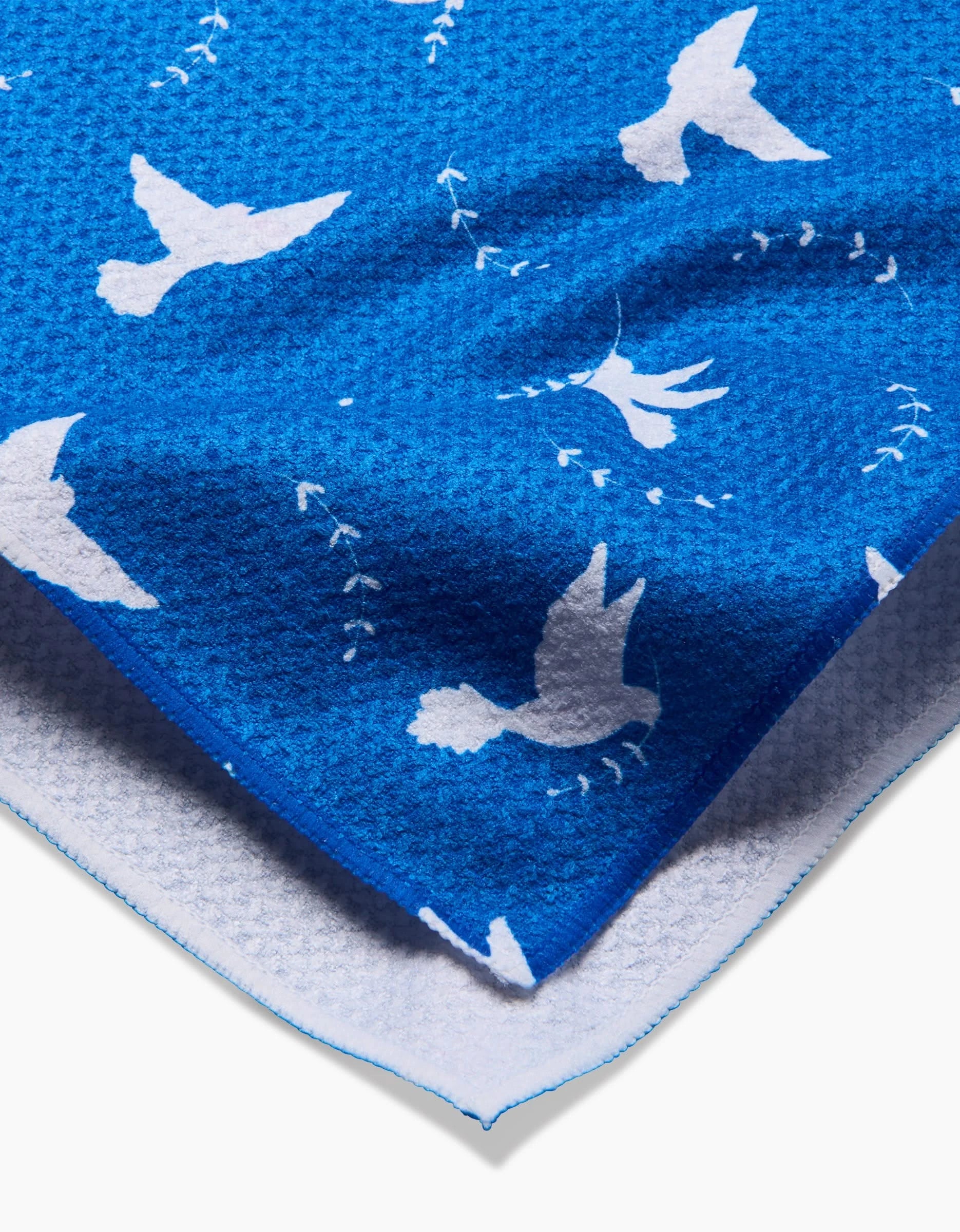 A close-up of the Peace Dove tea towel by Pascale Dilger, showing blue fabric with white doves and leaf patterns. The eco-friendly towel’s edge reveals a soft white underside.