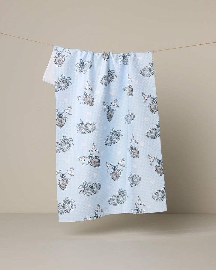 The Peace Doves kitchen tea towel by Geometry, crafted from recycled materials, features pale blue fabric adorned with hearts, ribbon-tied bundles, and white polka dot doves—all set against a beige background for an eco-friendly look.