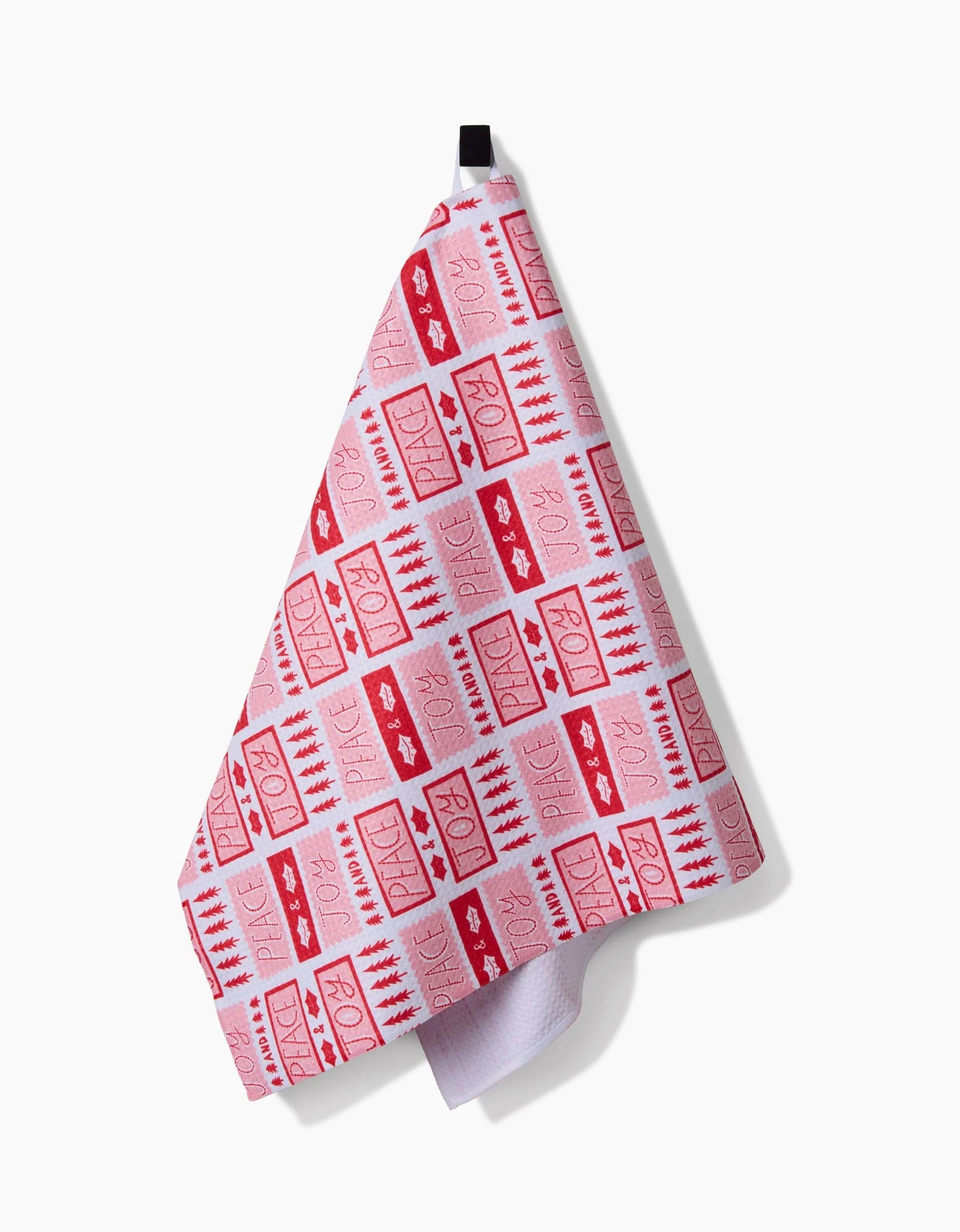 A tea towel from Roxy Marjs Peace Joy Stamps collection hangs from a black loop. Made from recycled materials, it showcases pink and red rectangular patterns on white, some stamped with Peace Joy and playful designs.