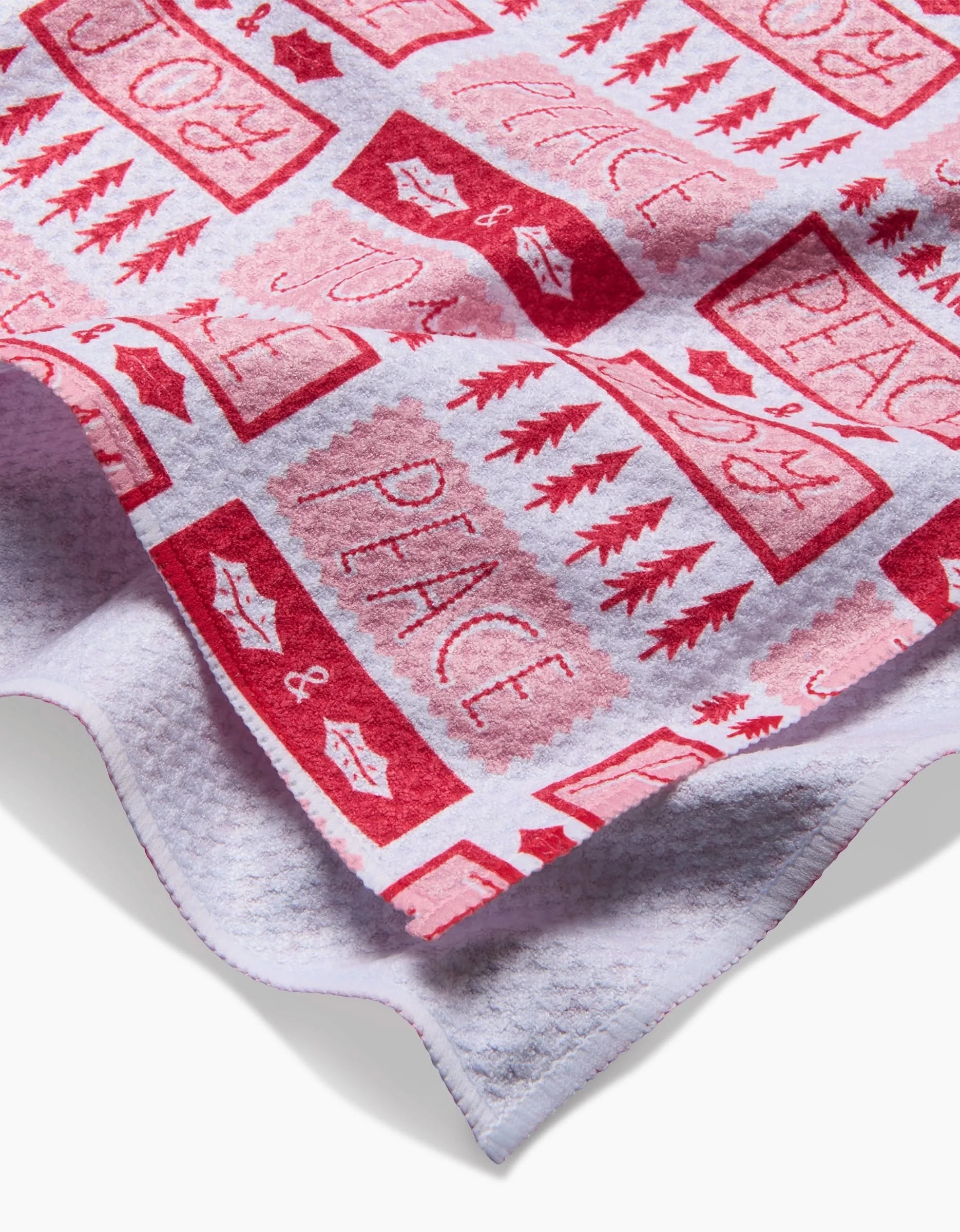 The Peace Joy Stamps tea towel by Roxy Marj features red and white festive patterns—trees, mittens, lips, and JOY & PEACE stamps—on recycled quick-drying fabric. The partially folded design highlights its textured surface.