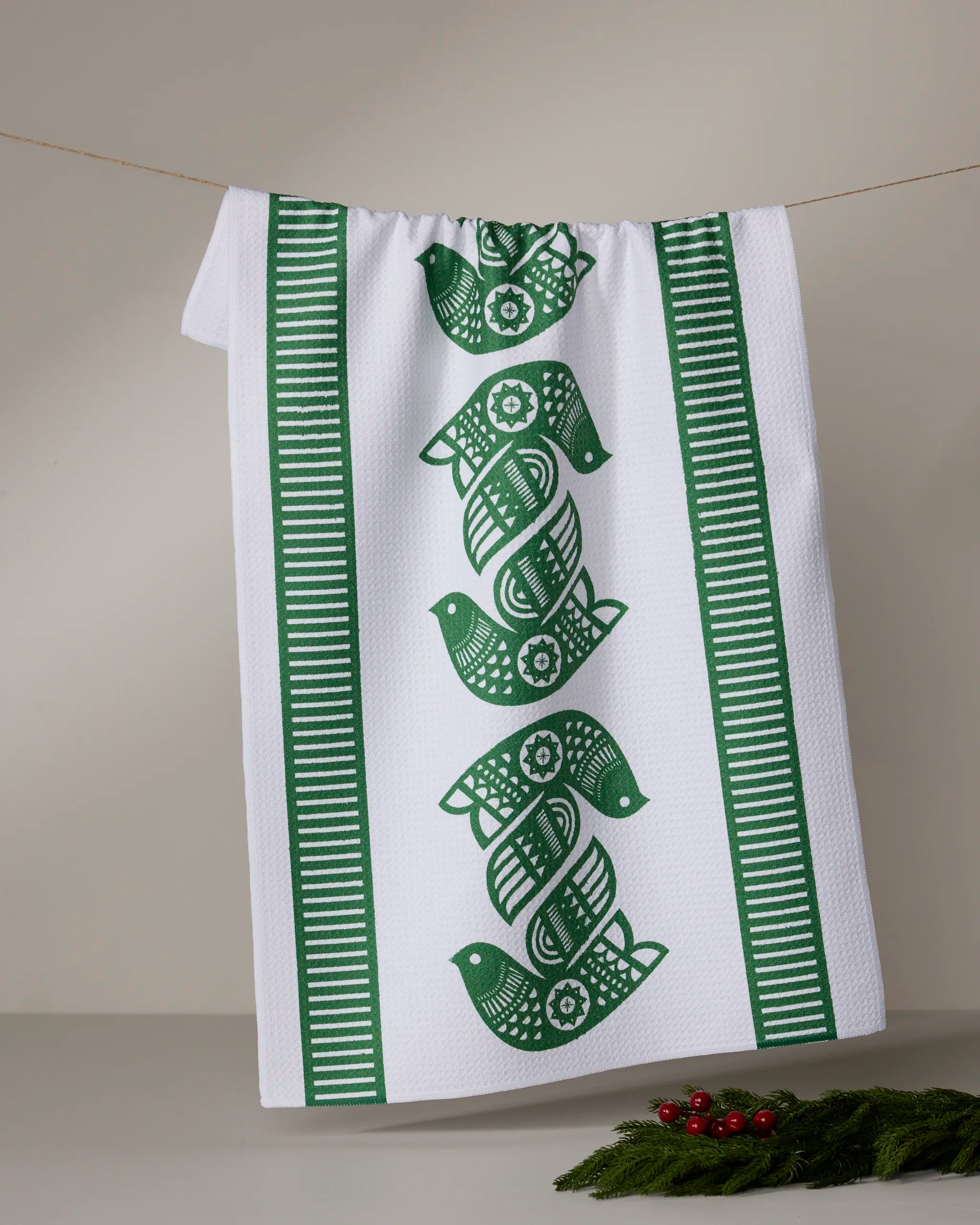 The Peacebound kitchen towel by Geometry, made from recycled materials, features green patterned birds stacked in the center with green and white striped borders. It hangs on a line, with evergreen sprigs and red berries nearby.
