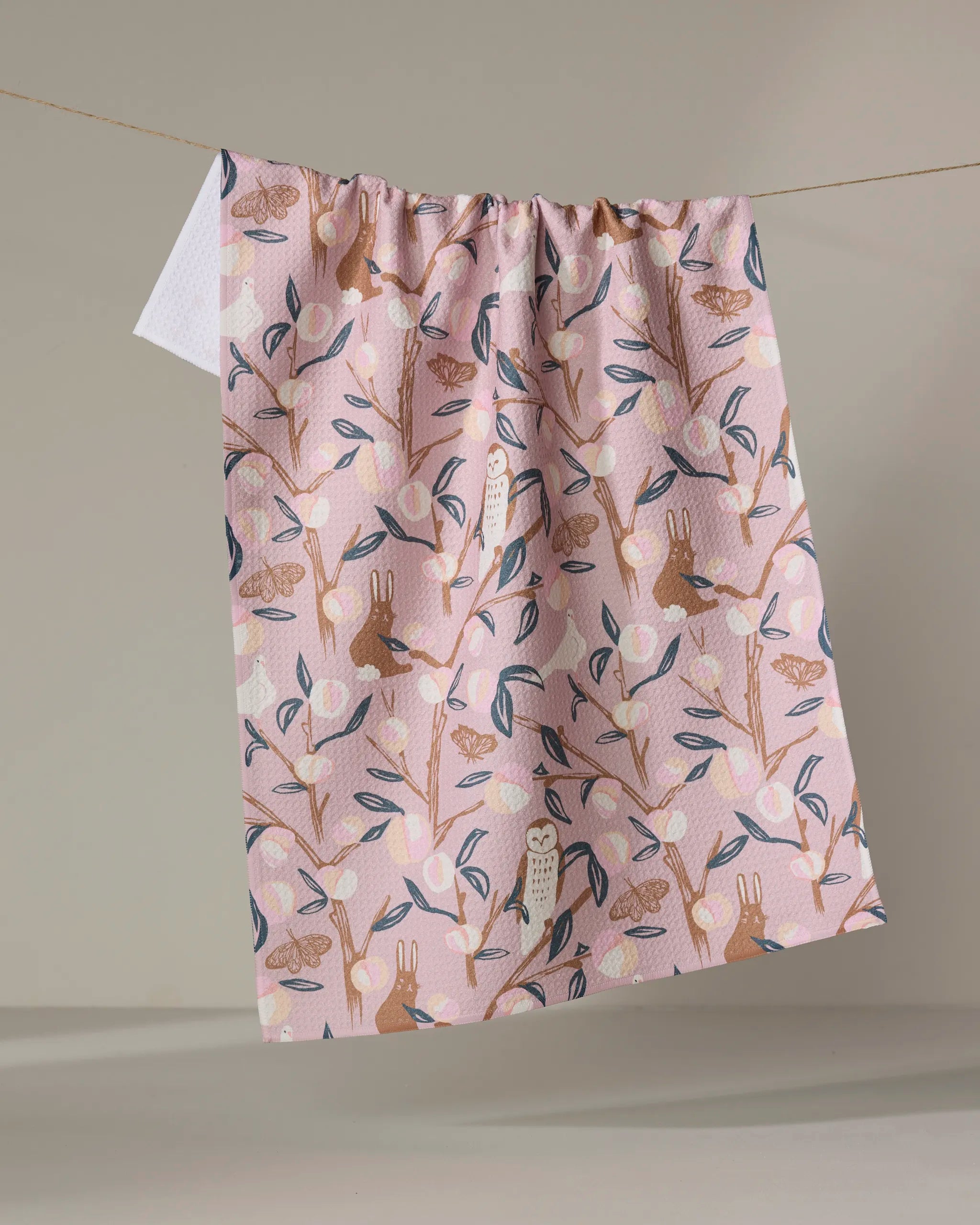 The Peach Tree tea towel by Elisabeth Wing features a charming owl, rabbit, and tree branch design in earthy tones on pink, hanging against a beige backdrop. This eco-friendly towel brings warmth and charm to your kitchen.