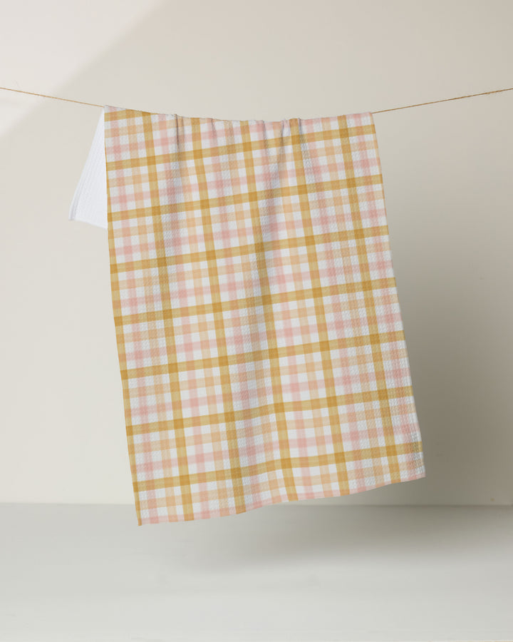 The Geometry Peachy Plaid tea towel, featuring a yellow, pink, and white pastel plaid design, hangs neatly on a string against a neutral background.
