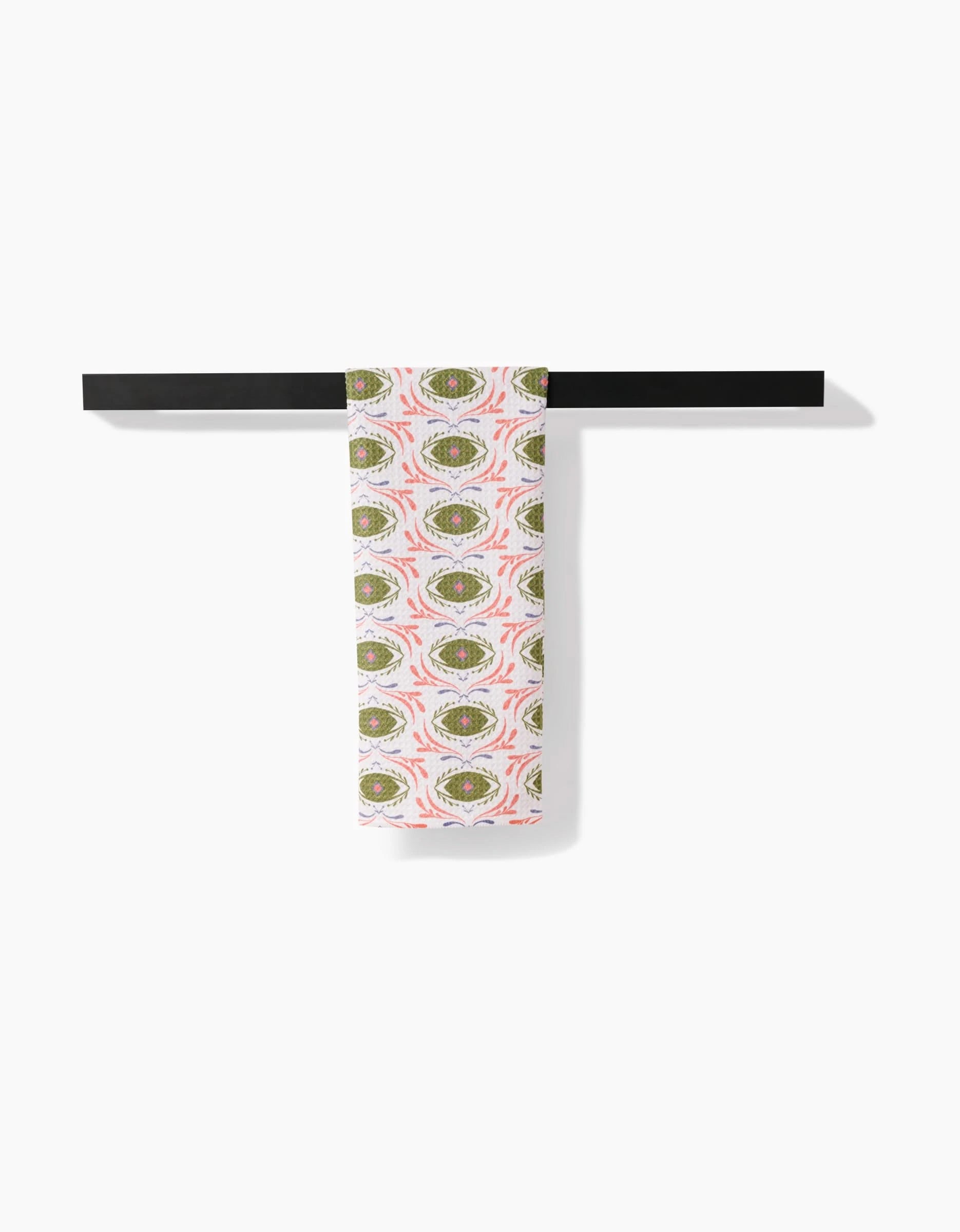 The “Peacock Eyes” tea towel by Amita Nair, featuring a green and pink geometric pattern, hangs neatly over a black bar against a plain white background.