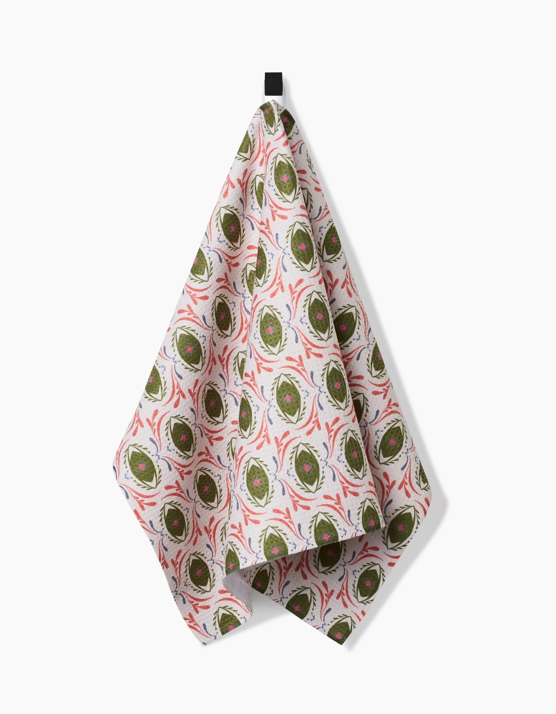The Peacock Eyes recycled kitchen towel by Amita Nair features a repeating pattern of sliced green and red vegetables and hangs from a black loop against a white background.