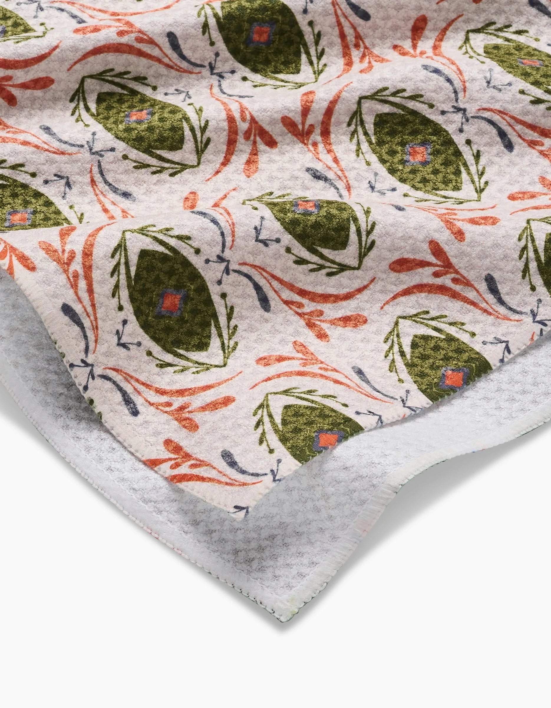 A close-up of the Peacock Eyes tea towel by Amita Nair, featuring green ovals with red and blue centers, abstract lines on a textured white background, and visible absorbent quick-drying edges in the corner.