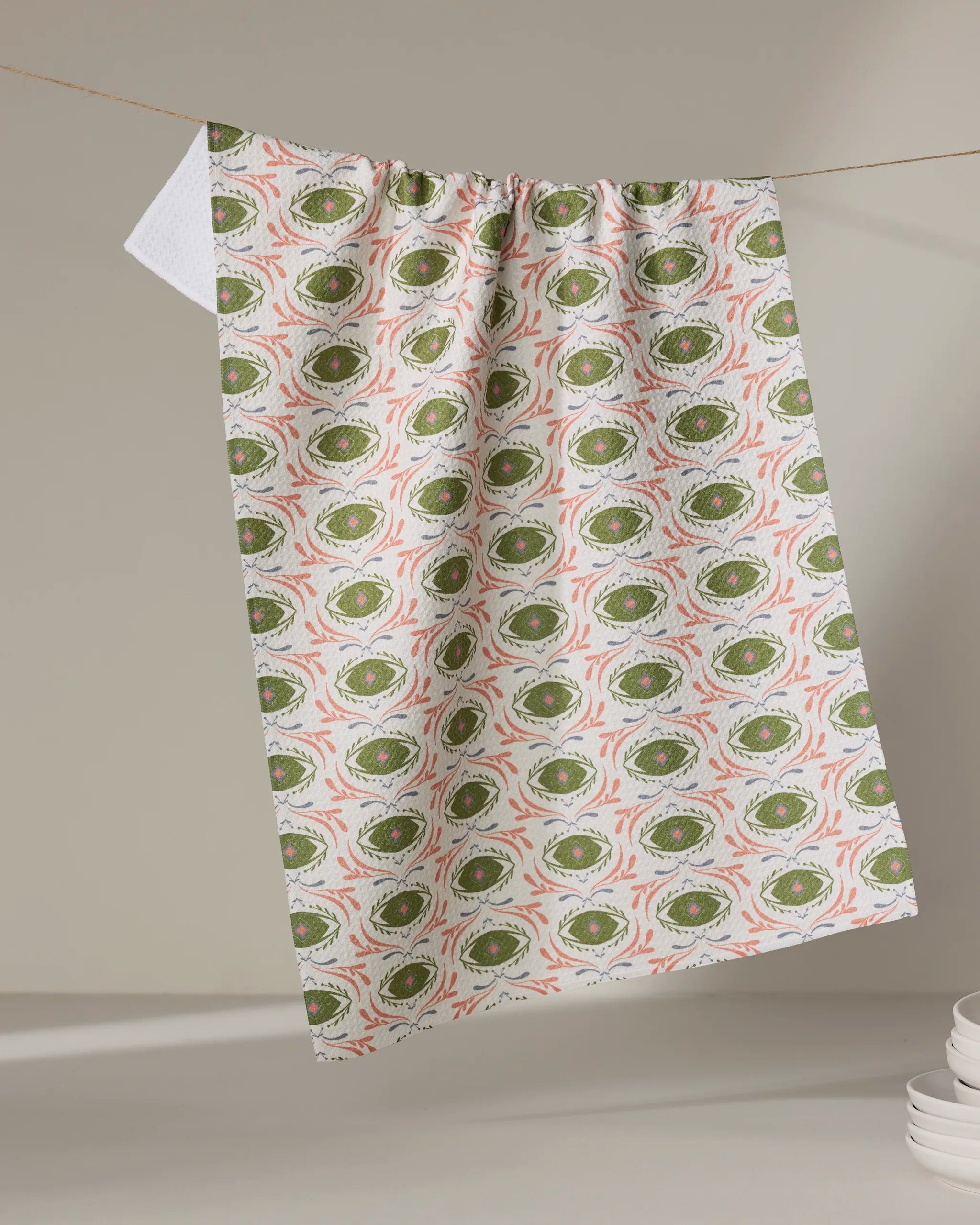 The "Peacock Eyes" kitchen towel by Amita Nair, featuring green and orange abstract designs, hangs from an indoor clothesline. Stacks of white plates appear at the lower right corner of the image.