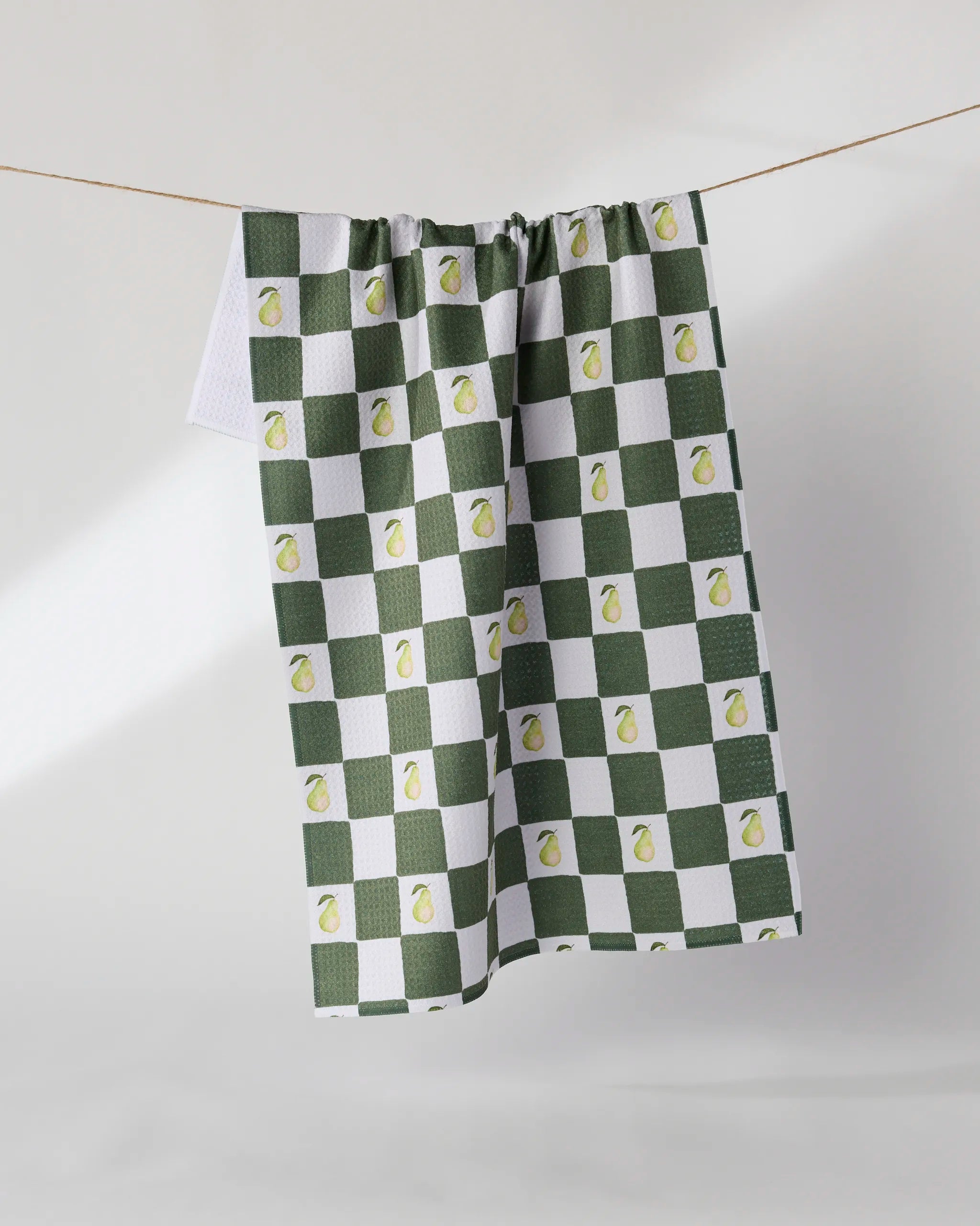 The Pear Checker tea towel by Geometry features a green and white checkerboard design with small pear illustrations, hangs on a clothesline against a light background, and is made from recycled materials for eco-friendly appeal.