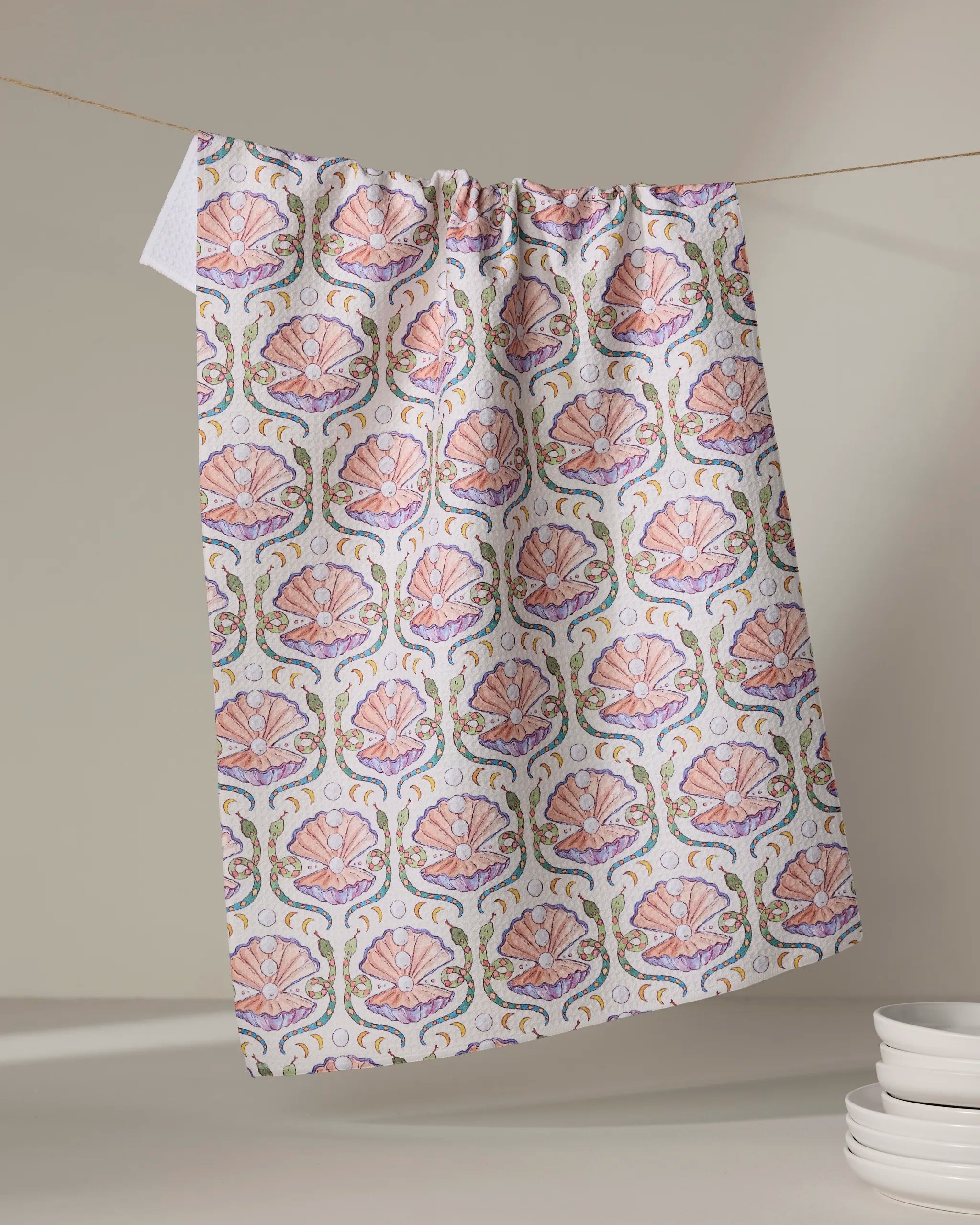 The Pearls Of The Moon kitchen towel by Maayan Alper-Swan, featuring a pink and green seashell and scroll design, hangs on a line with a neutral background and a stack of white dishes in the lower right corner.