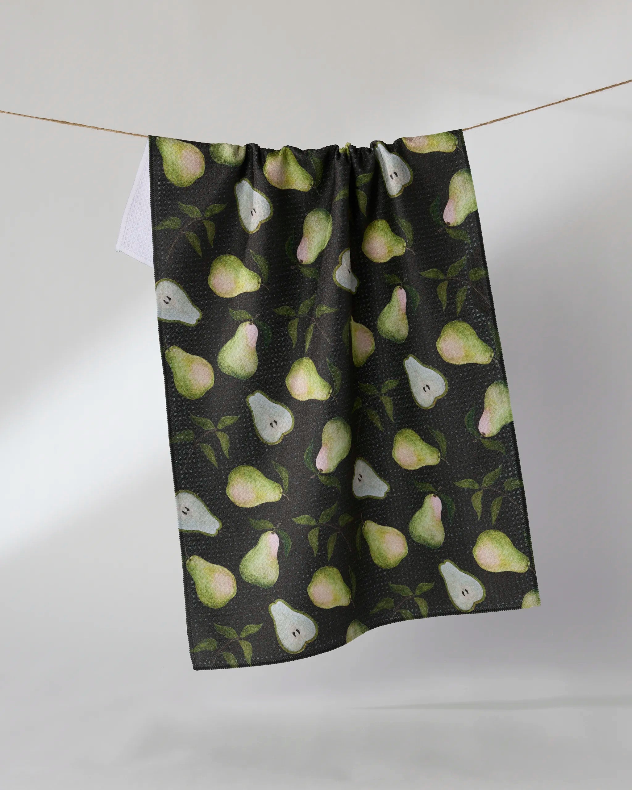 The Geometry Pearwood eco-friendly tea towel, featuring green and yellow pears and leaves on a dark background, hangs on a clothesline against a plain light wall.