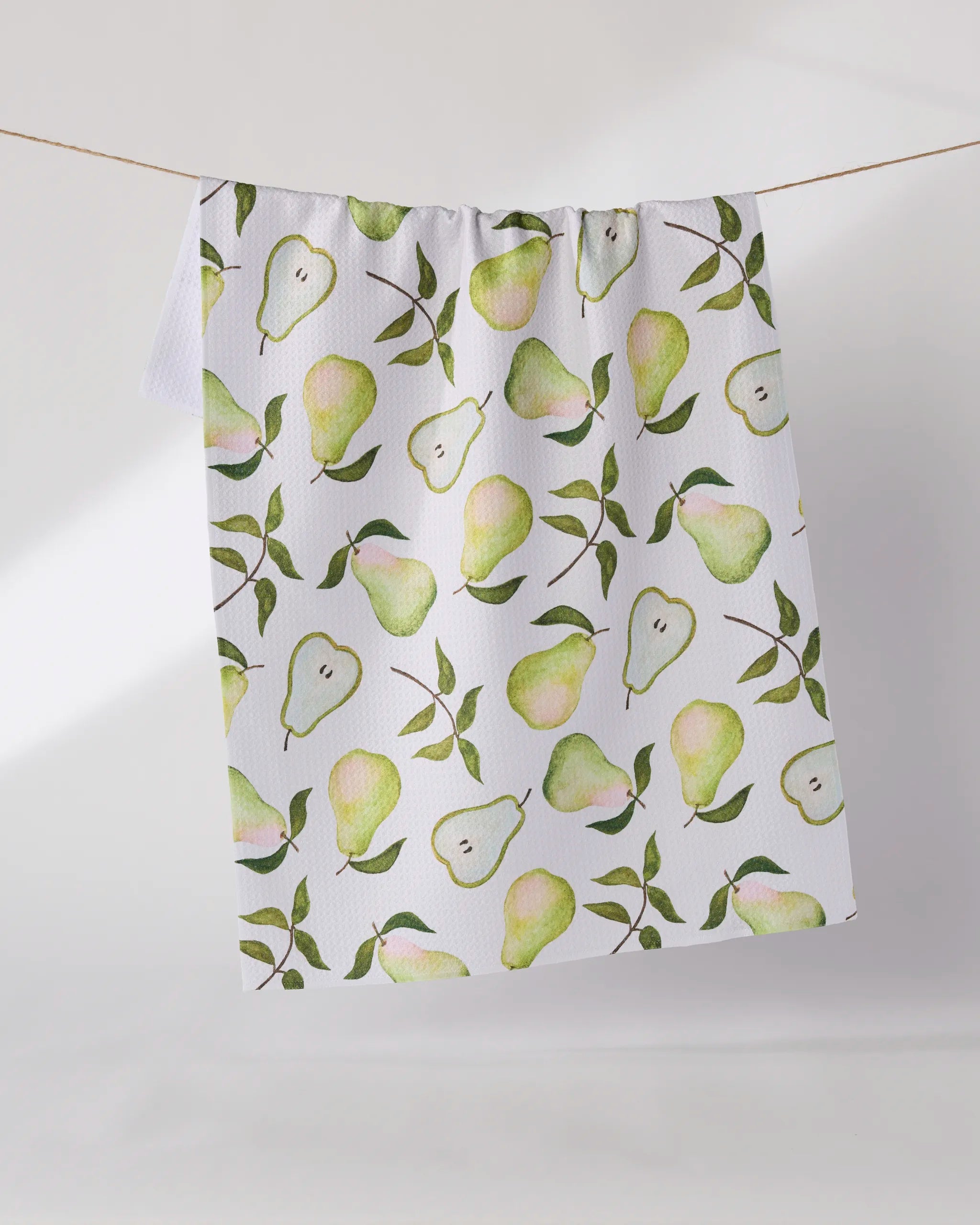The Geometry Pearwood tea towel, in white with a watercolor-style pear and green leaf pattern, hangs on a line against a light background.