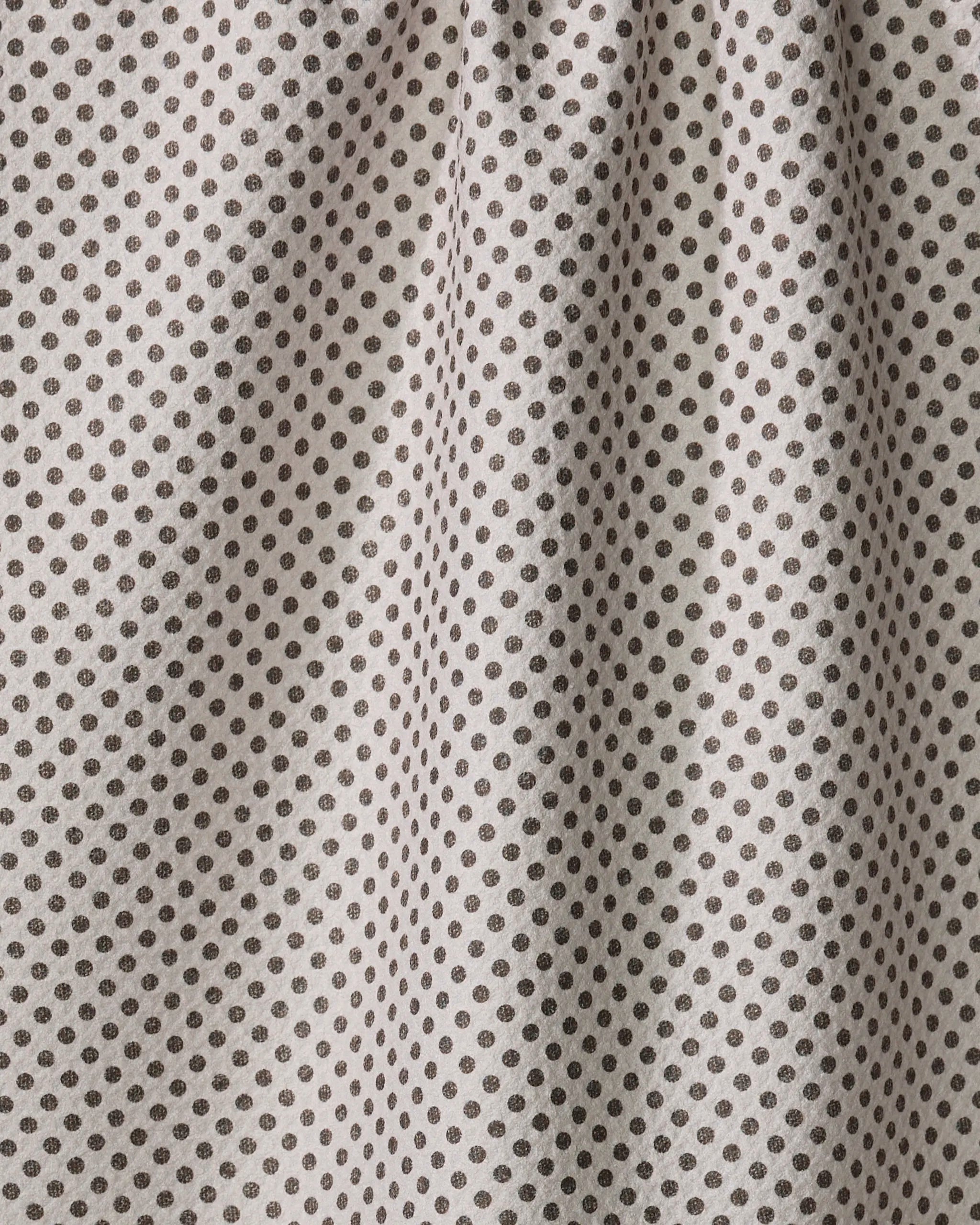 Close-up of the Pebble tea towel by Jenny Keller, crafted from beige fabric with evenly spaced small black polka dots. Soft vertical folds and shadows add texture and charm to this practical kitchen essential.