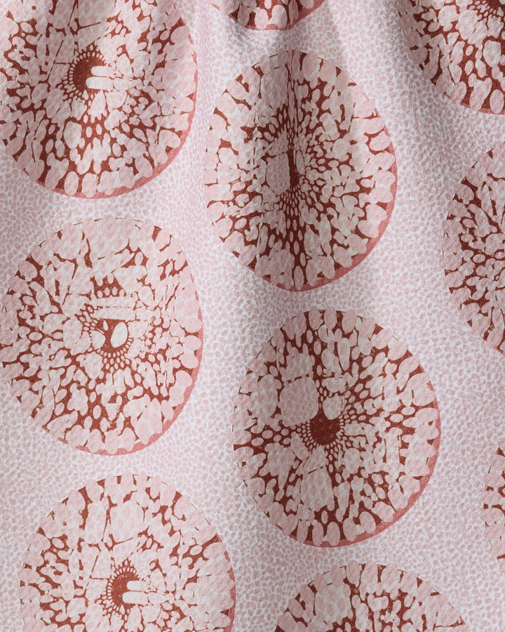 A close-up of the Dabney Lee “Pebbles” tea towel shows circular, reddish-brown patterns like cell structures or tree rings on light pink fabric, highlighting its subtle folds and texture.