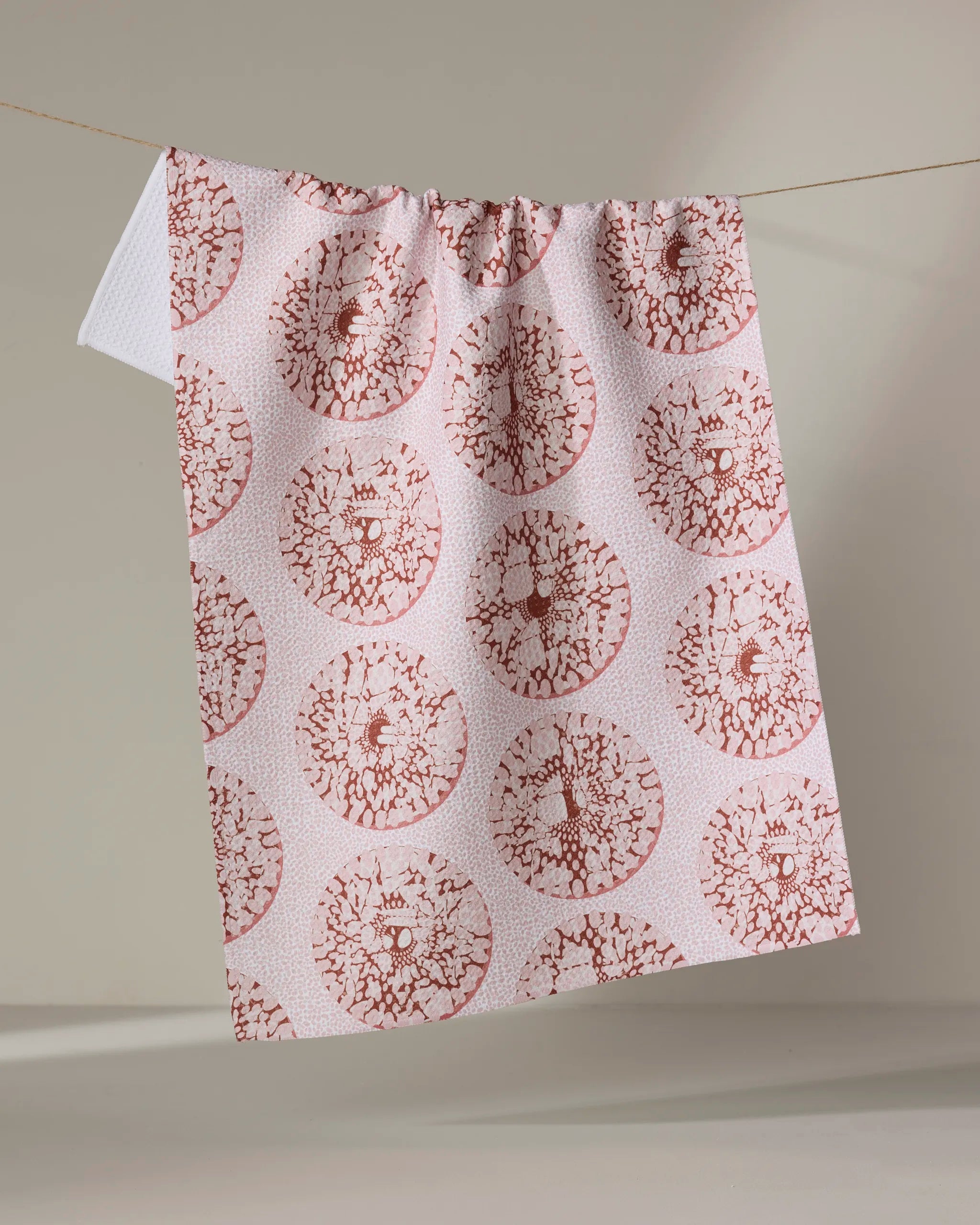 The "Pebbles" kitchen towel by Dabney Lee, featuring red circular floral patterns and made from recycled materials, hangs on a string against a neutral background.