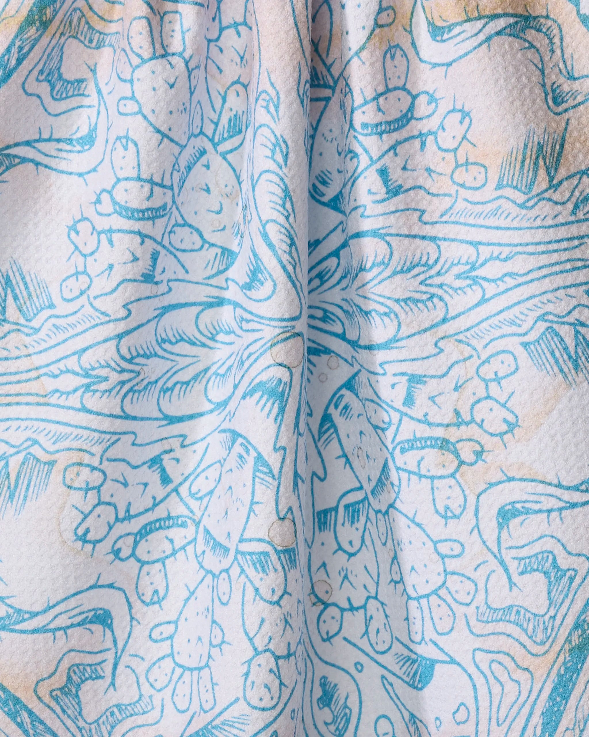 A close-up of the Pelican tea towel by Skye Walker, made from recycled materials with a blue symmetrical pattern of abstract shapes, cartoon cacti, mushrooms, and plants, slightly gathered at the center.