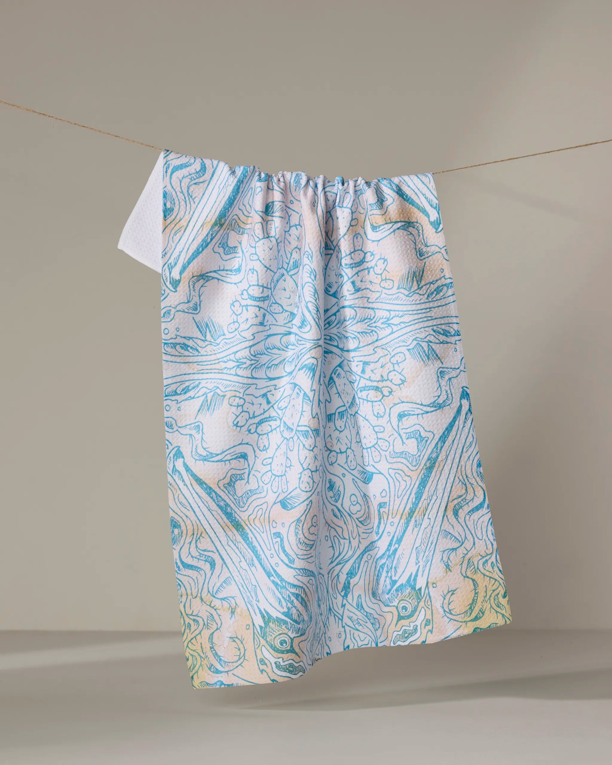 The Skye Walker "Pelican" kitchen towel, featuring an intricate marbled pattern in blue, white, and yellow pastels, hangs on a clothesline against a plain background.