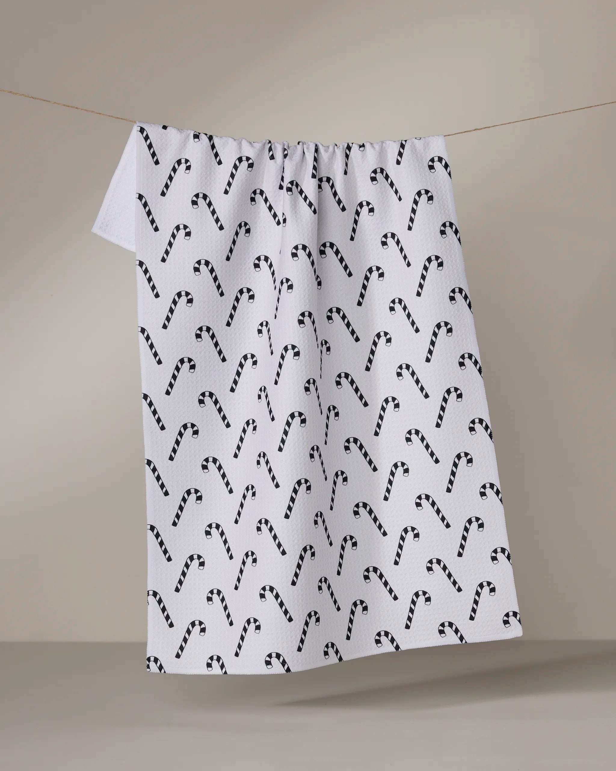 The Geometry Peppermint Pop towel, a quick-drying white cloth with a black candy cane pattern, hangs from a string against a plain, neutral background.