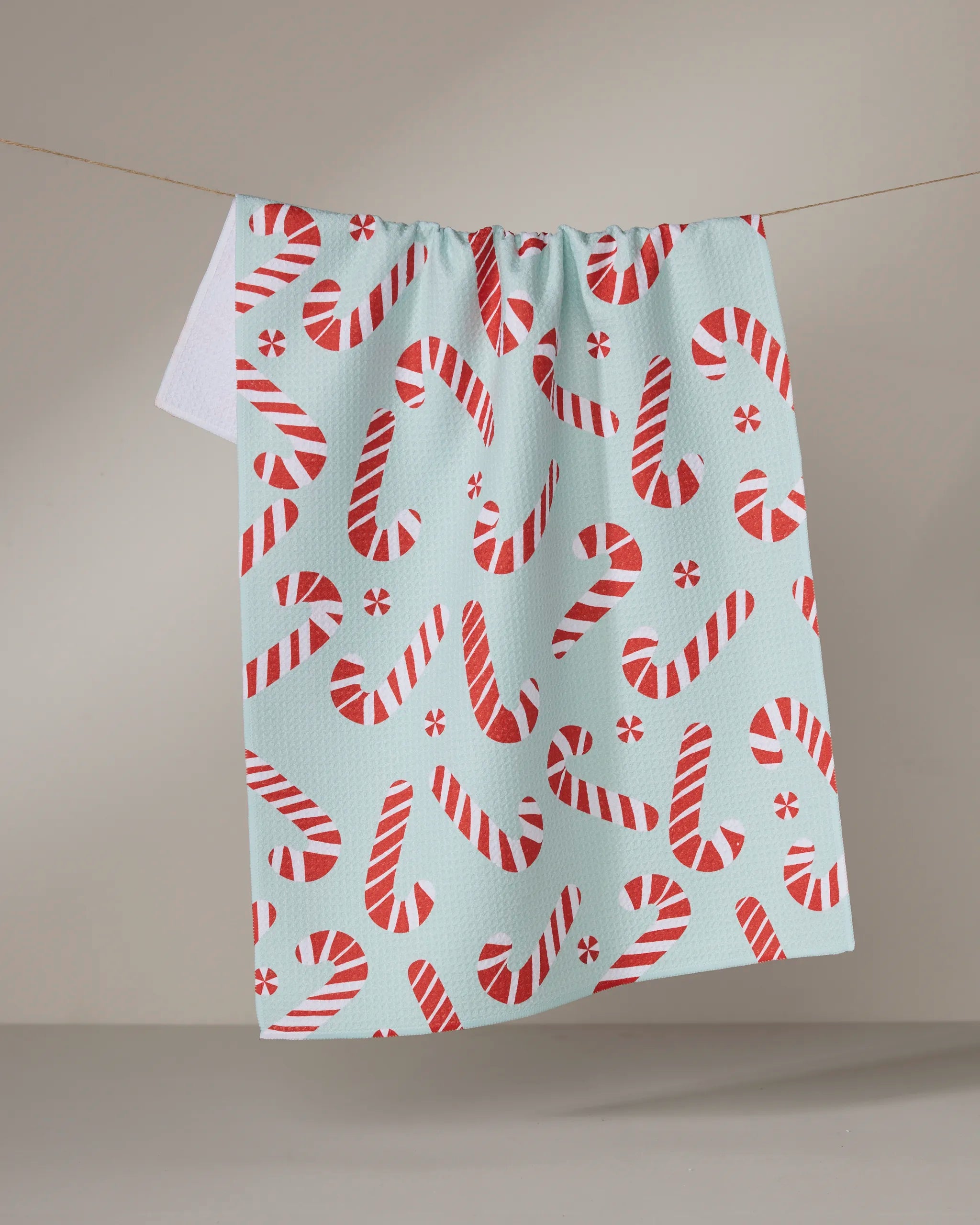 The Peppermint Twist recycled kitchen towel by Geometry, featuring a red and white candy cane pattern, hangs on a clothesline against a plain, light background.