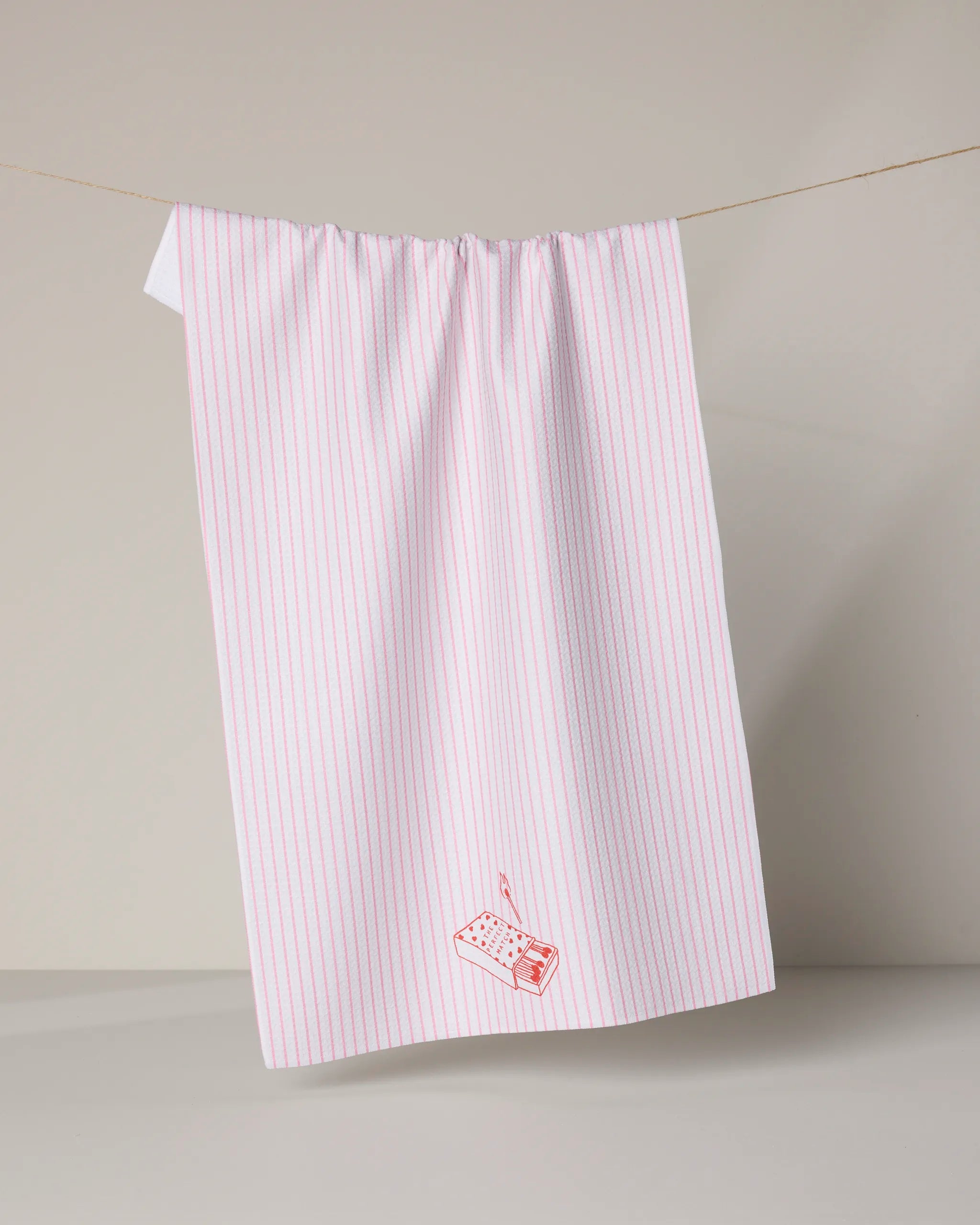Geometry's Perfect Match tea towel, made from recycled materials, hangs on a line. It features thin red vertical stripes and a small radio illustration near the bottom corner against a plain neutral background.