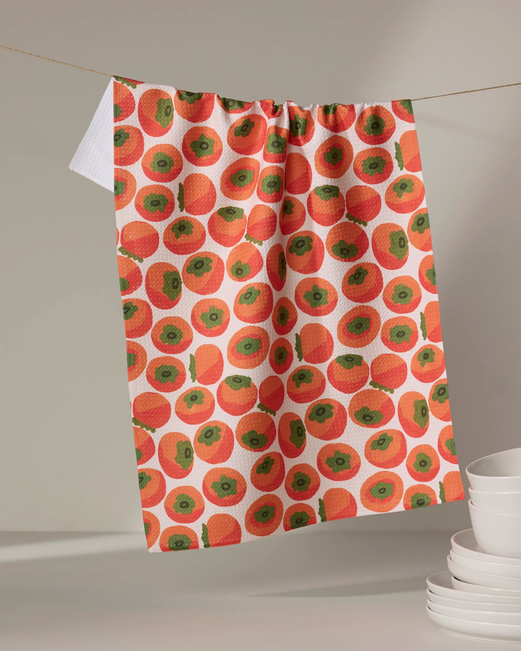 The Persimmons kitchen towel by Katie O'Shea, featuring a vibrant pattern of red and green circles on white, hangs on a clothesline with a stack of white bowls nearby against a neutral backdrop.