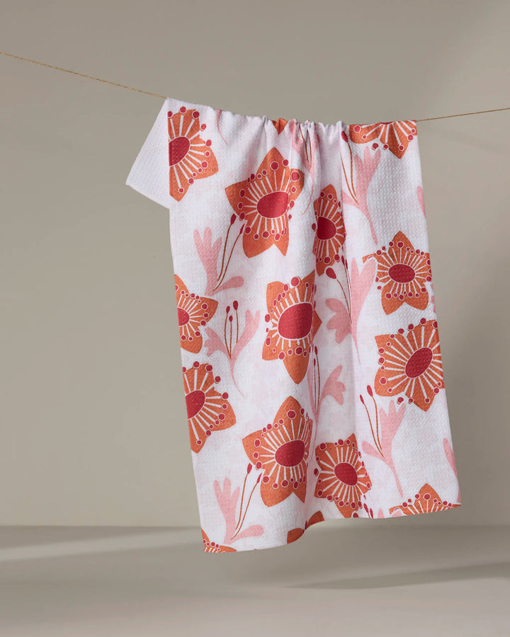 The Petal Paradise recycled kitchen towel by Whitney Urquhart, featuring a red and pink floral pattern, hangs on a clothesline against a neutral background.