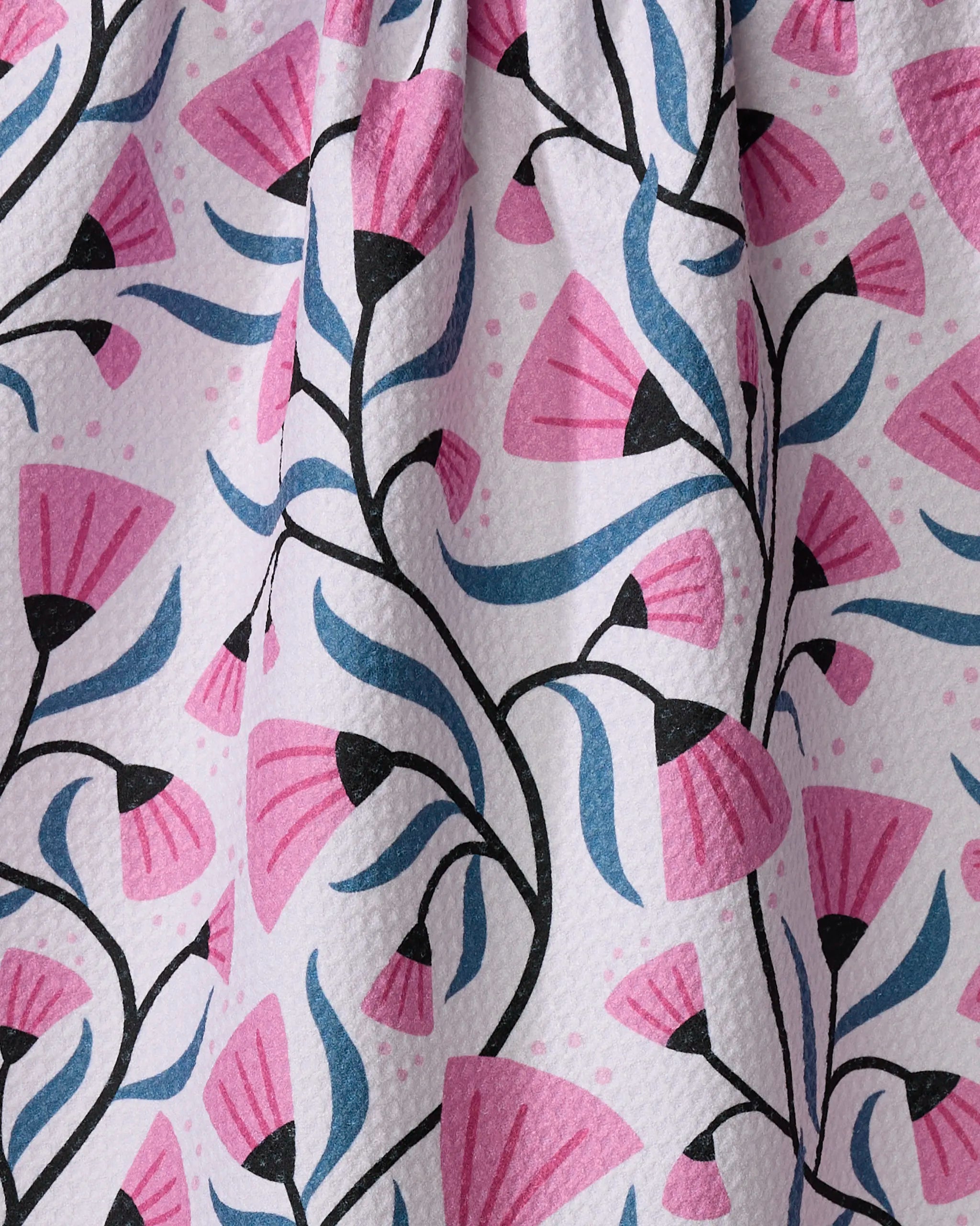 Close-up of Anette Heiberg's Petal Symphony dish towel with pink floral patterns, blue leaves, and black stems on a white textured background. The absorbent fabric is gently draped with soft folds.