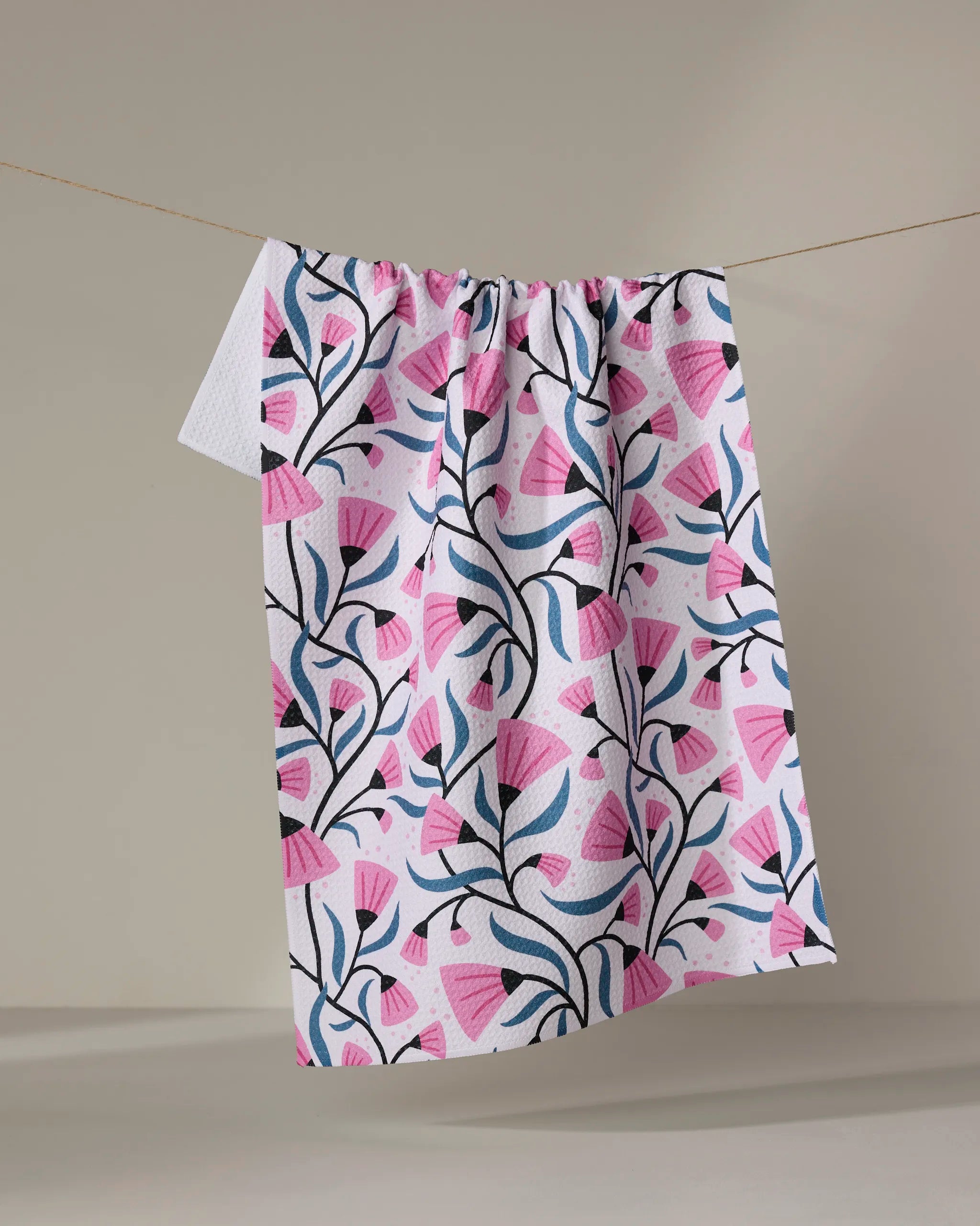 The Anette Heiberg "Petal Symphony" dish towel, featuring a pink and blue floral pattern, hangs on a clothesline against a neutral background.