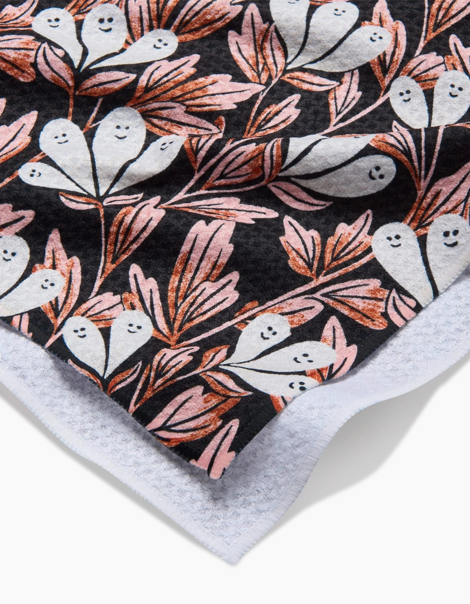 A close-up of the Sarah Daugherty Phantom Floral ultra-absorbent towel, showing small, smiling white ghosts with pink and orange leaves on a black background and its white terry cloth edge in the foreground.