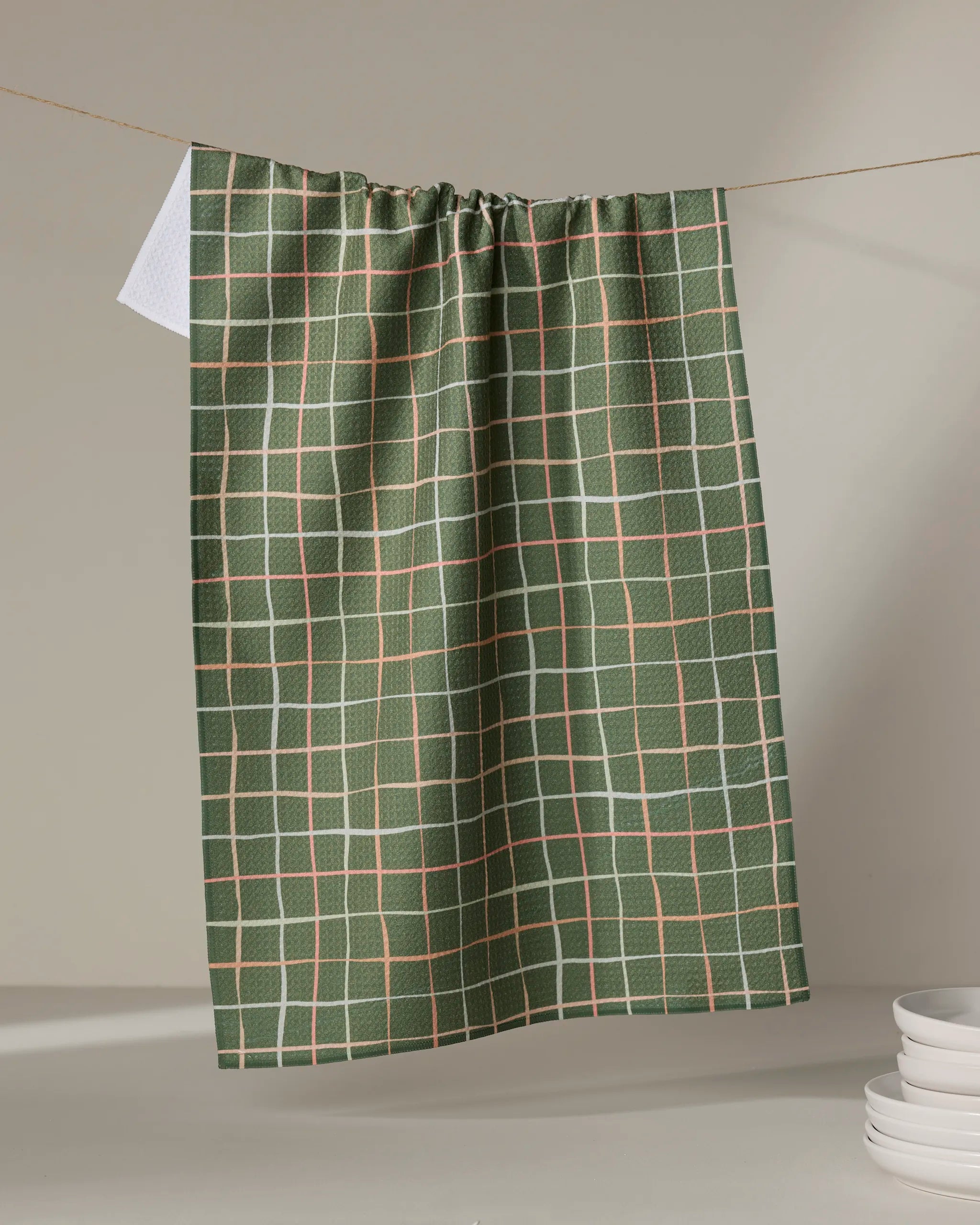 The "Picnic" tea towel by Anee Shah, featuring a green checkered pattern with white and tan lines and made from recycled materials, hangs on a string. White dishes are stacked in the lower right against a neutral background.