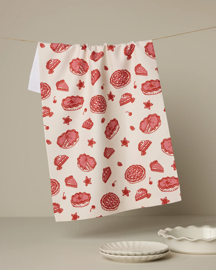 The Geometry Pie-triotic recycled kitchen towel, featuring a red and pink pie and star pattern, hangs above a beige surface next to stacked white plates and an empty pie dish.