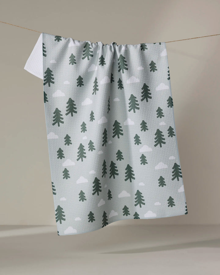 The Pine Forest kitchen tea towel by Coit Creative, featuring green pine trees and white clouds, is made from recycled materials and hangs on a clothesline against a neutral background.